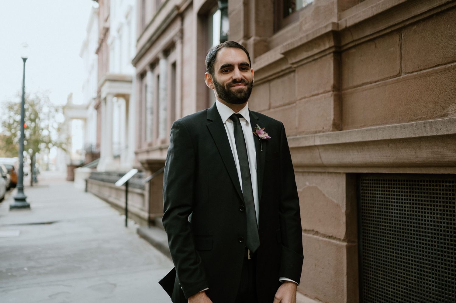 Groom in dark suit and tie stands against brownstone building exterior.