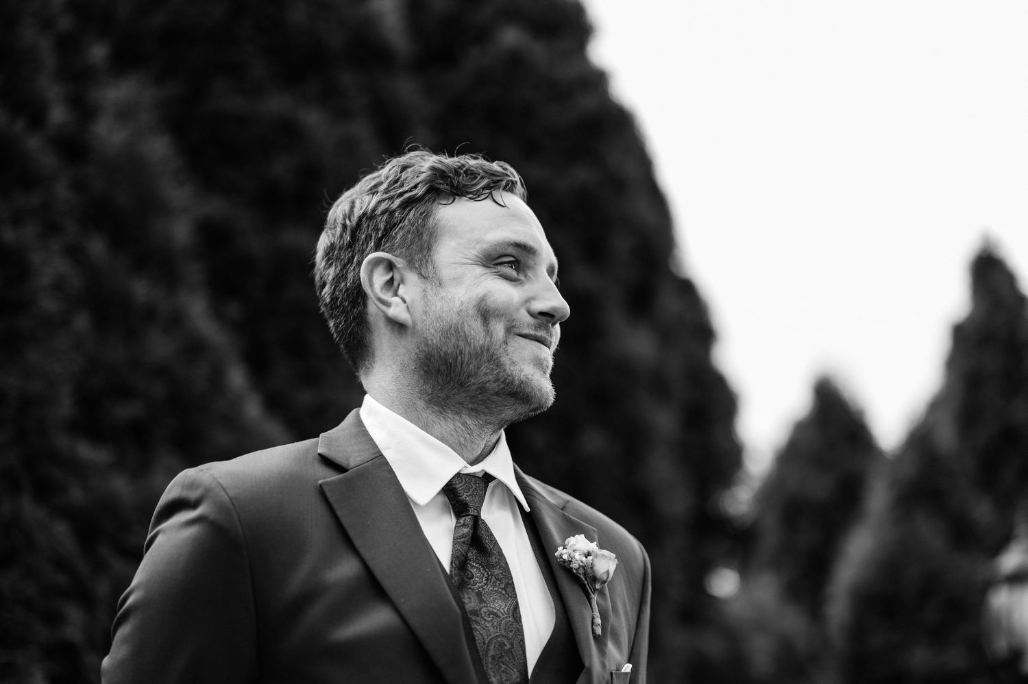 Black and white portrait of a groom smiling against a backdrop of trees.