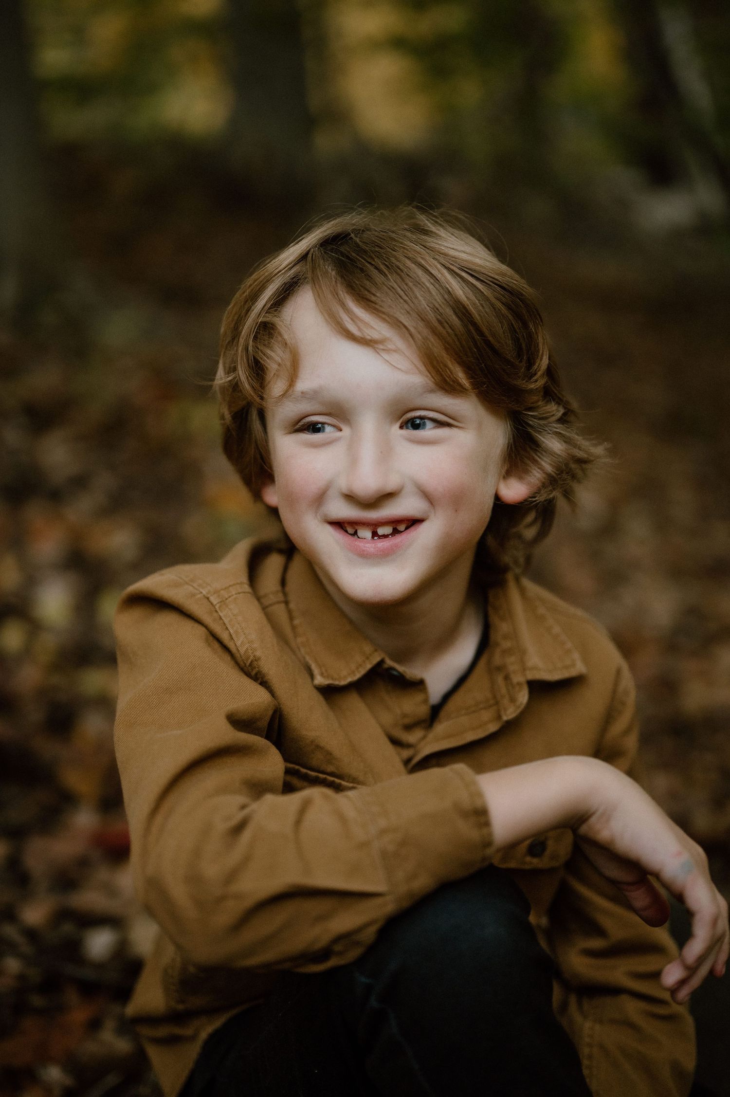 A joyful portrait of a child wearing a brown jacket sitting outdoors among autumn leaves.
