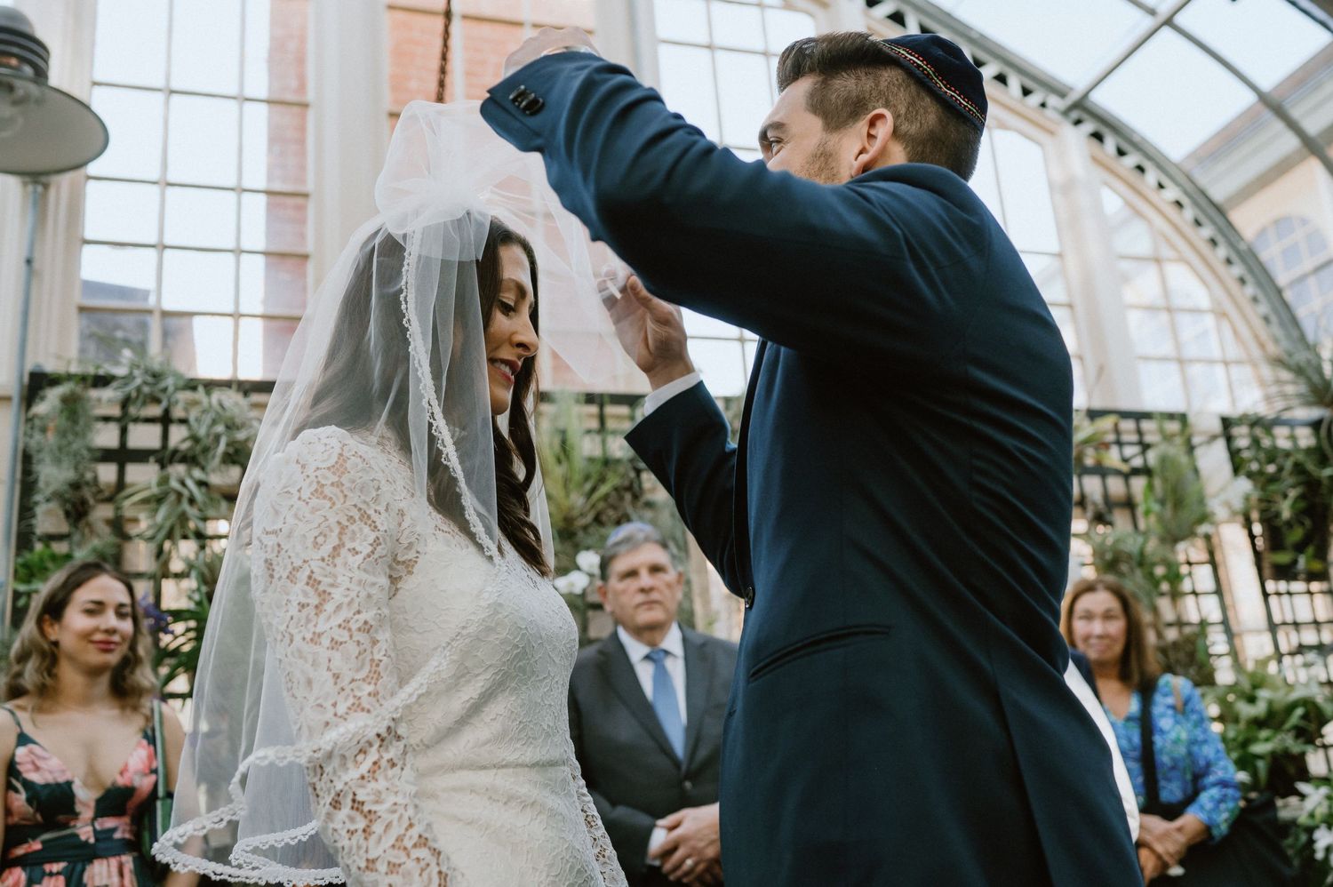 A wedding ceremony takes place in a beautiful greenhouse setting with natural light streaming through.