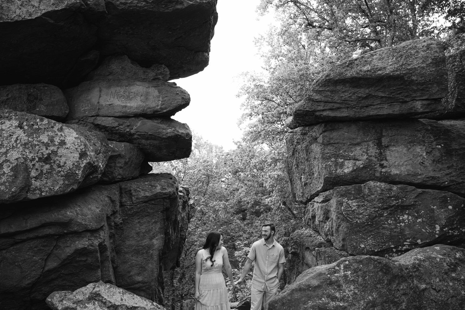 Couple holds hands walking between large rock formations in a black and white outdoor engagement photo.