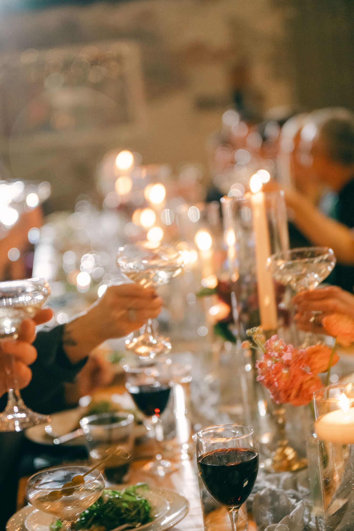 Guests raising crystal glasses in a candlelit dinner toast, with red wine and floral centerpieces on the table.