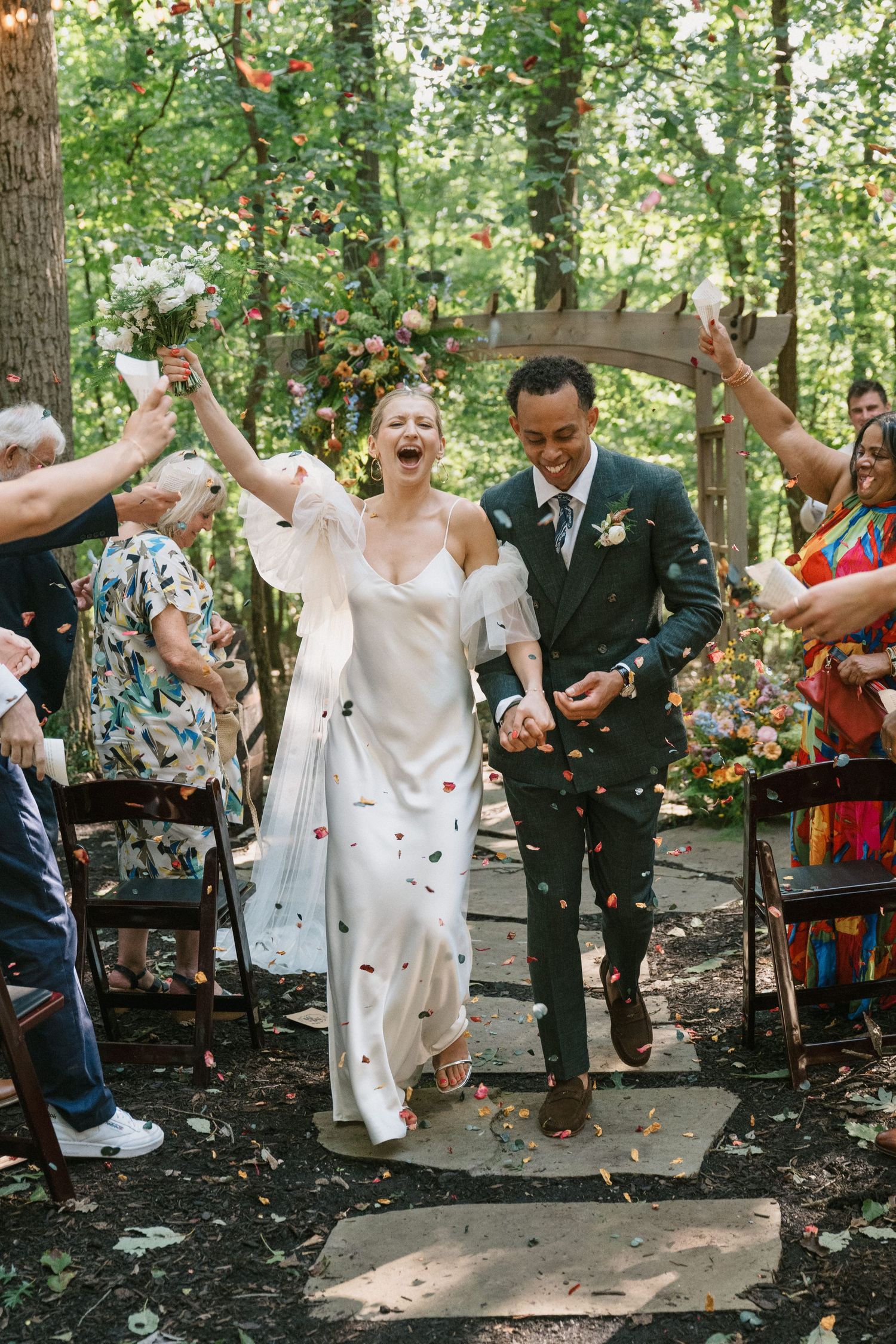Wedding celebration with guests throwing confetti in an outdoor forest setting.