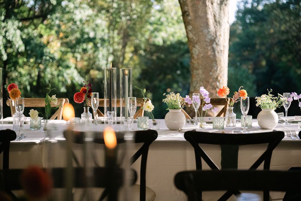 Elegant outdoor reception tables set under trees with white tent canopies.