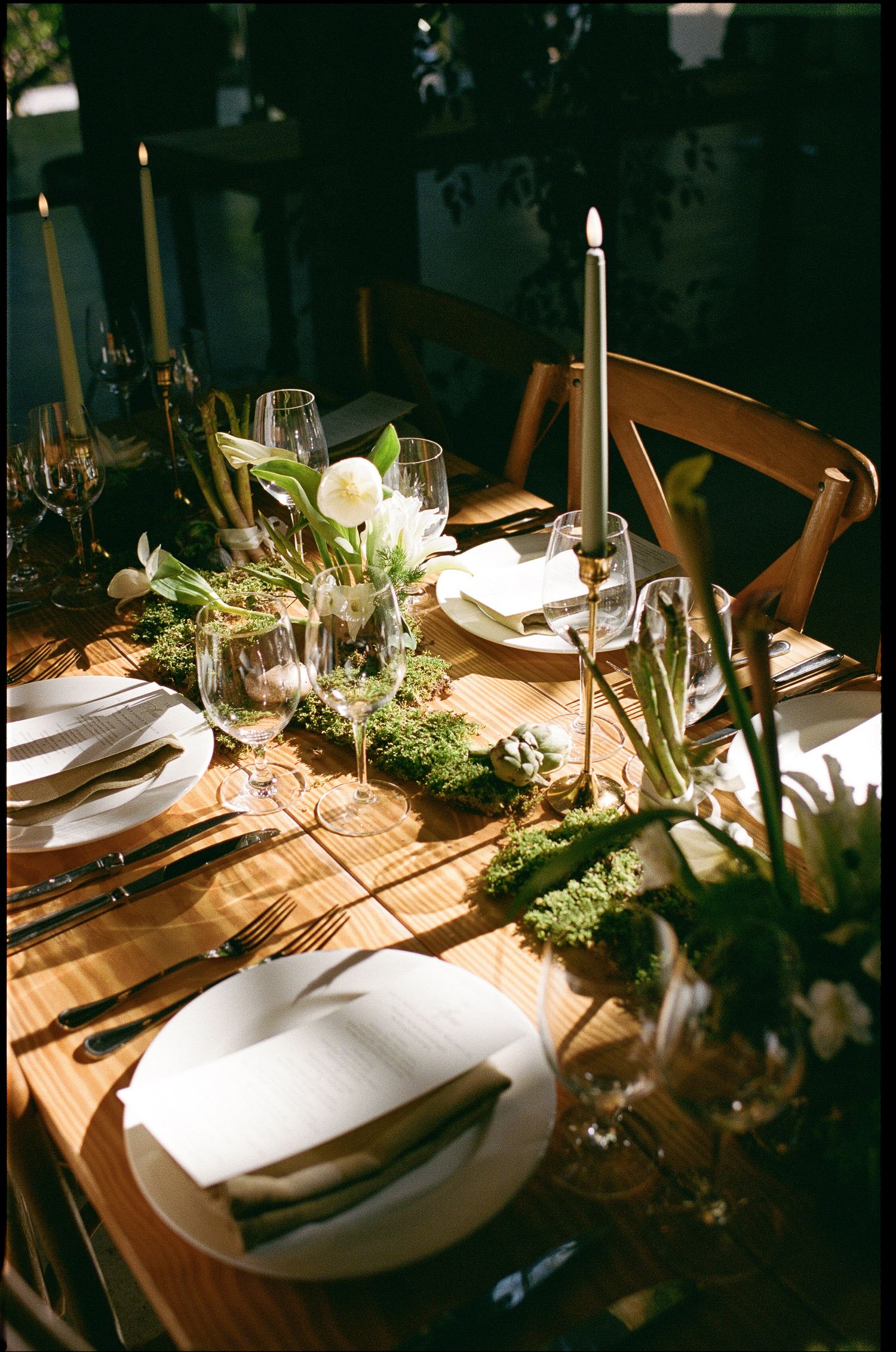 Elegant wooden dining table set with white plates, crystal glasses, green moss runner, white tulips, and tall taper candles.