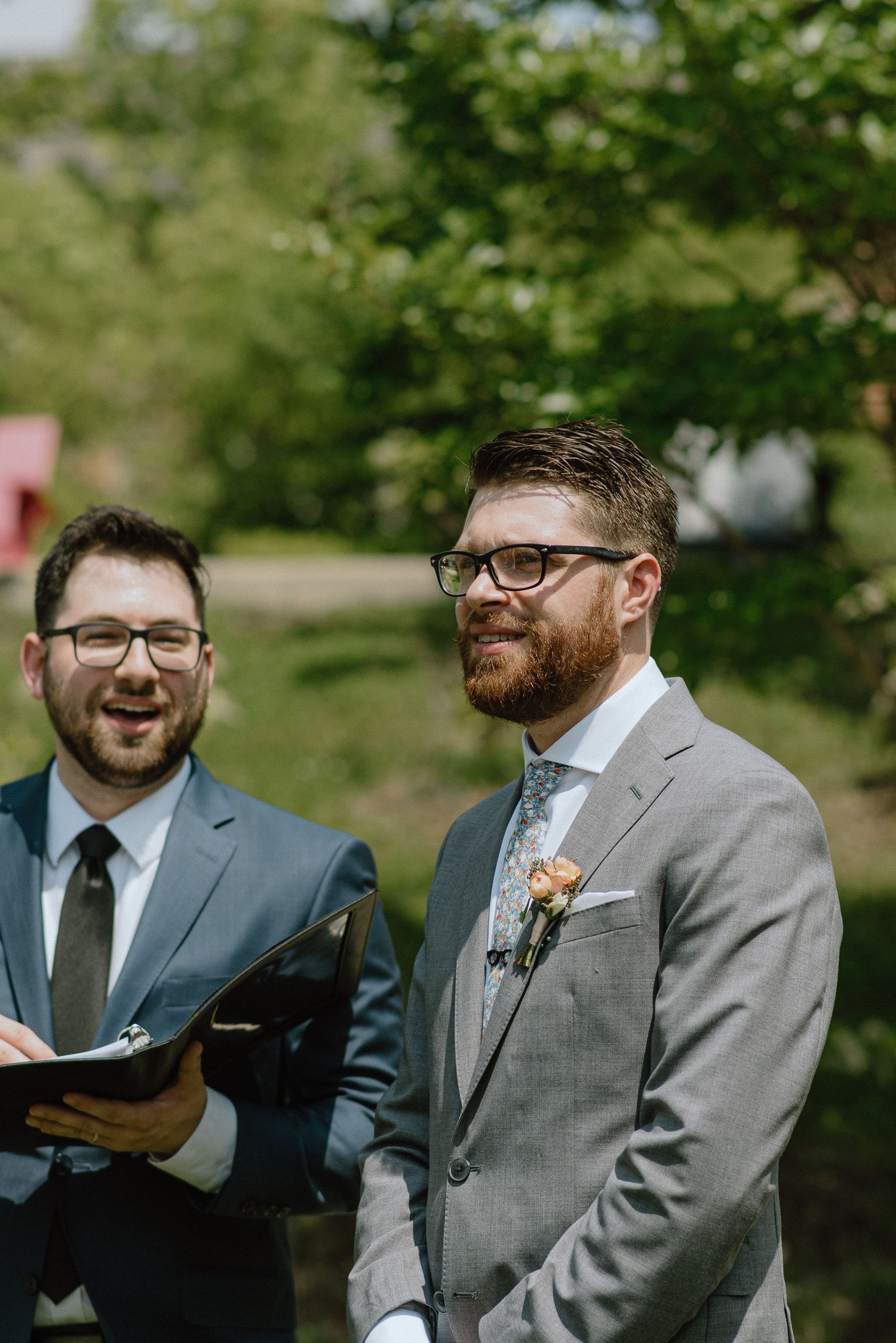 Two men in grey suits and glasses sharing a moment during outdoor ceremony.