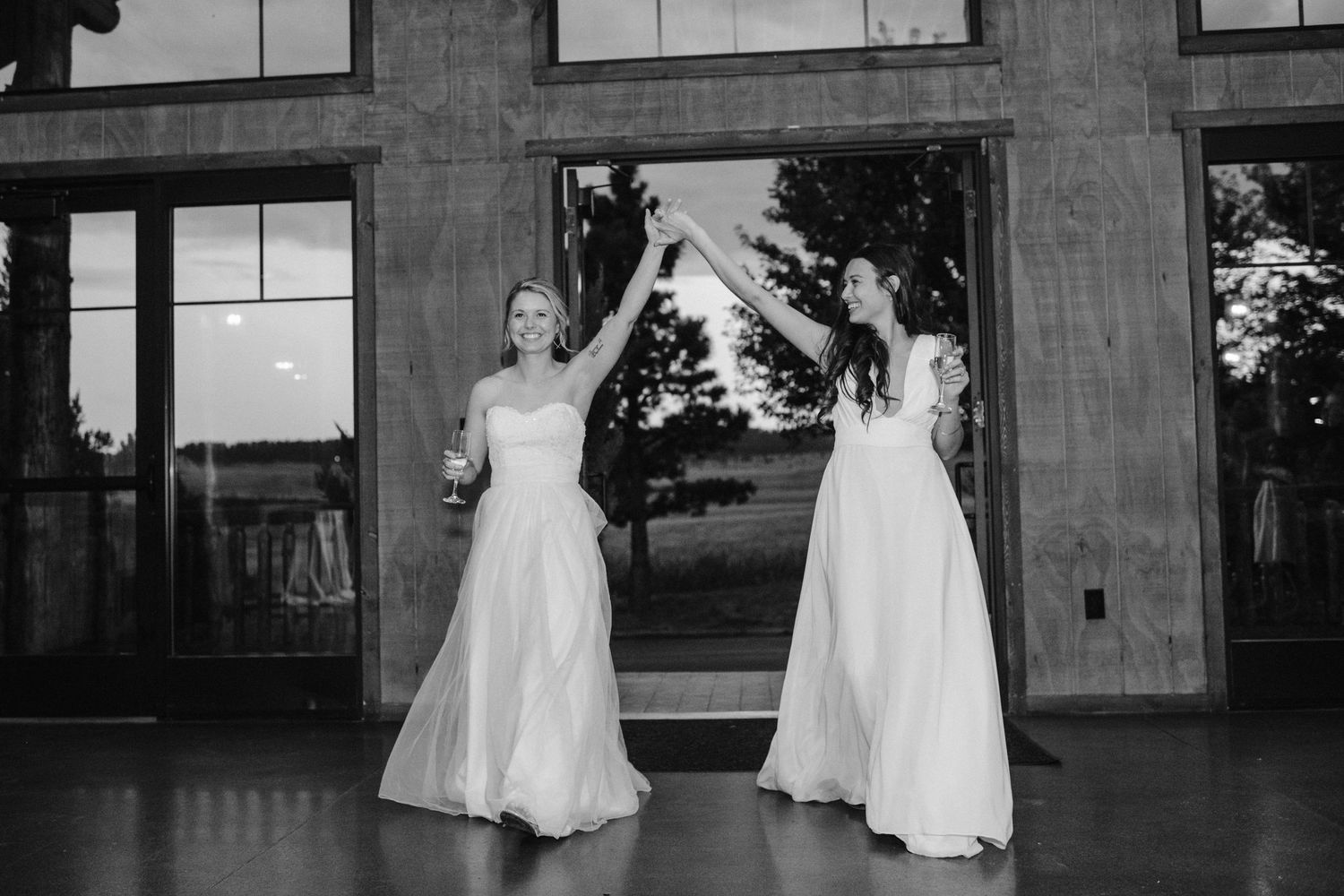 Two people in white wedding dresses dance together in front of large windows.