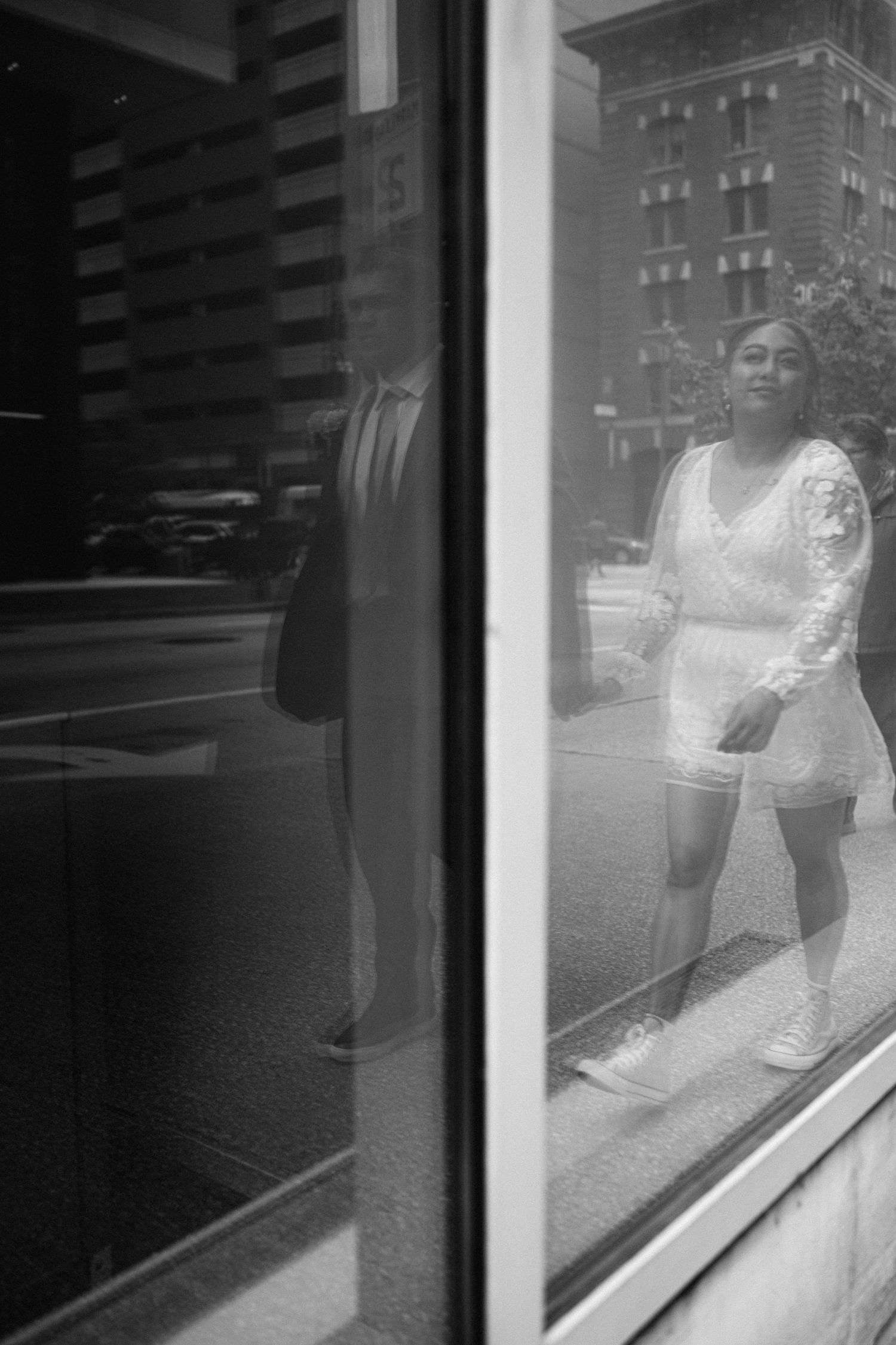 A black and white reflection through a window shows a person in a white romper standing on a city street.