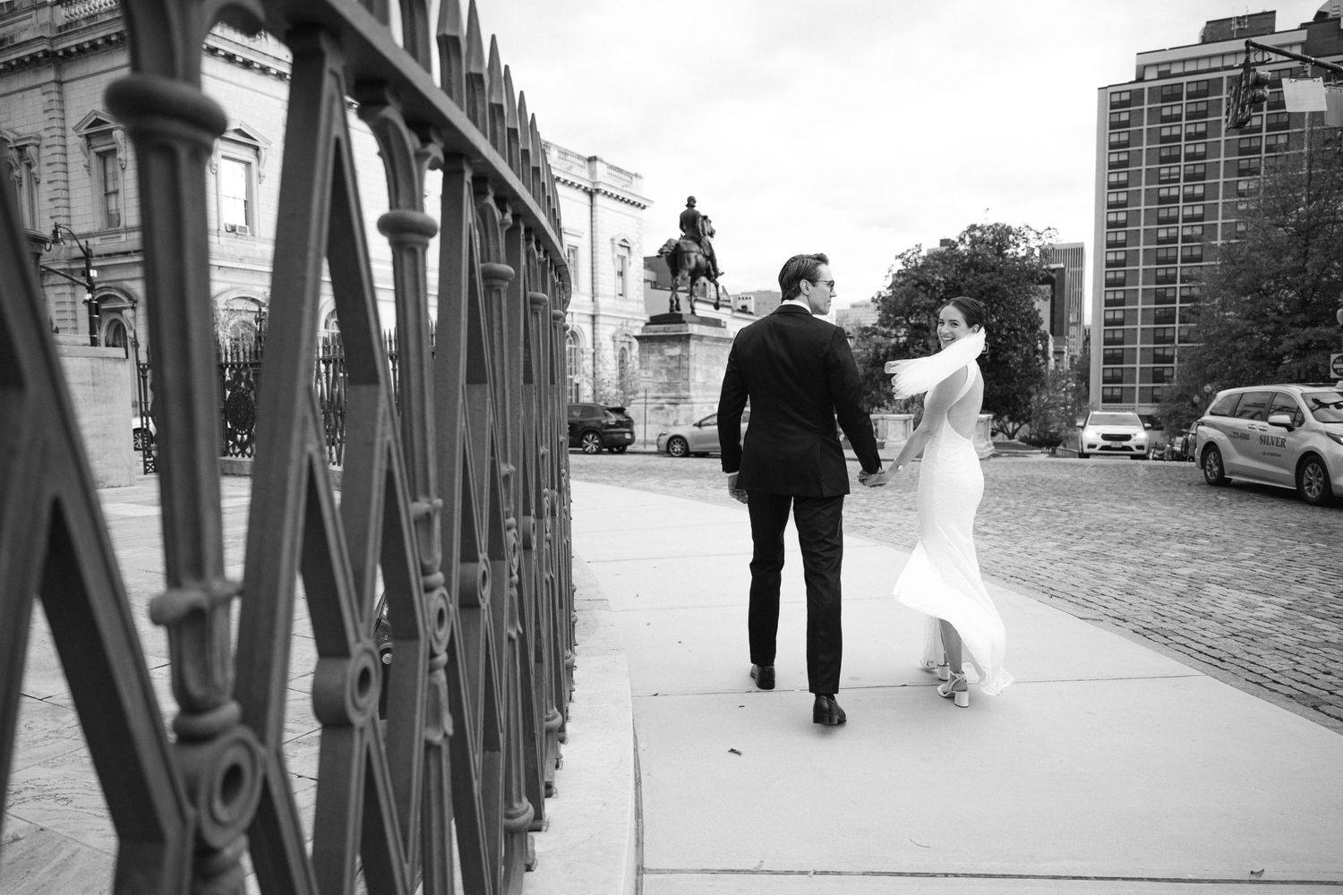 Black and white photo of newlyweds walking together on a bridge with cityscape in background.