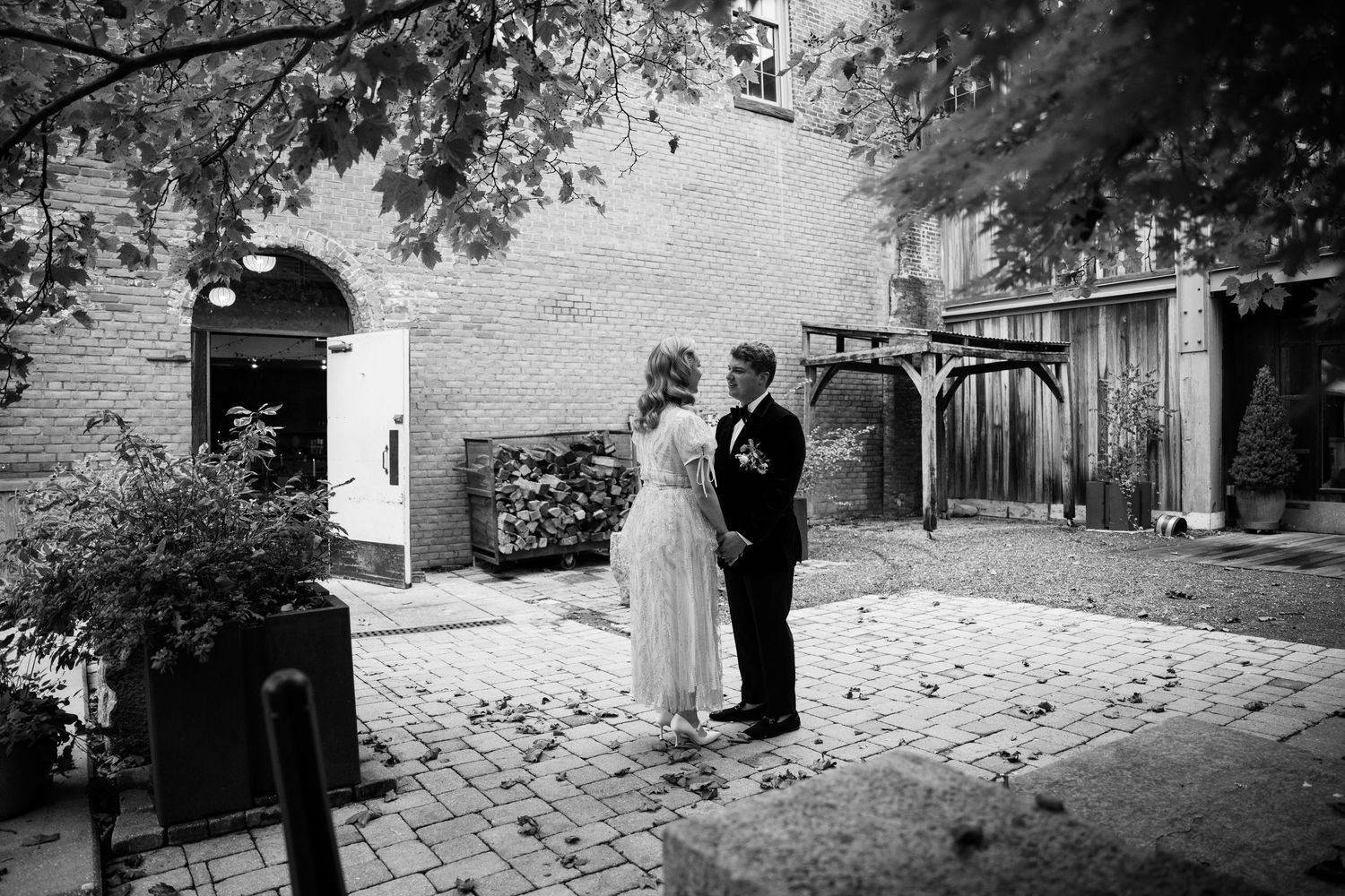 A black and white wedding photo shows a couple standing on brick pavement under tree branches.