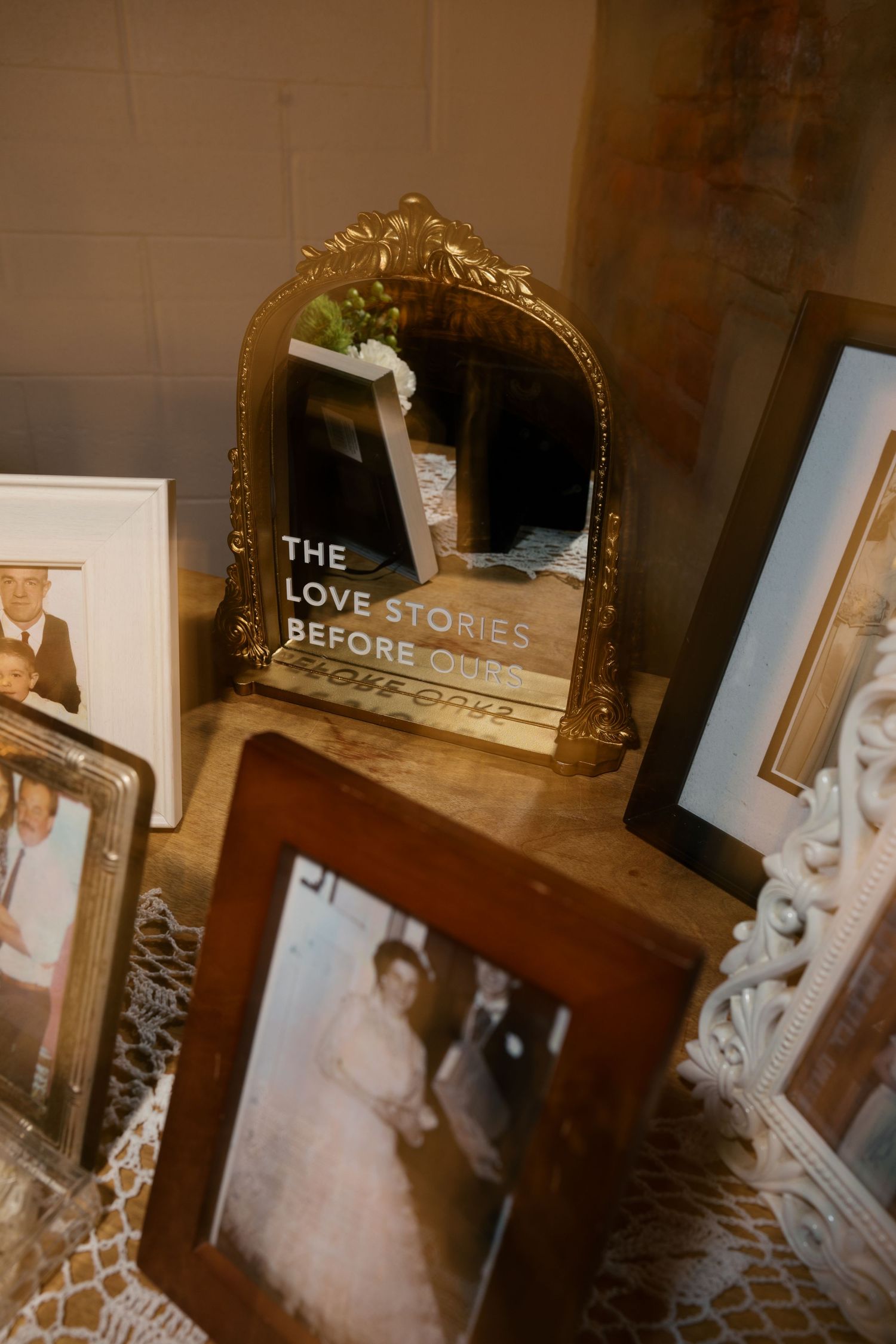 A vintage gold ornate mirror reflects a book titled 'The Love Stories Before' surrounded by framed photos and decor.