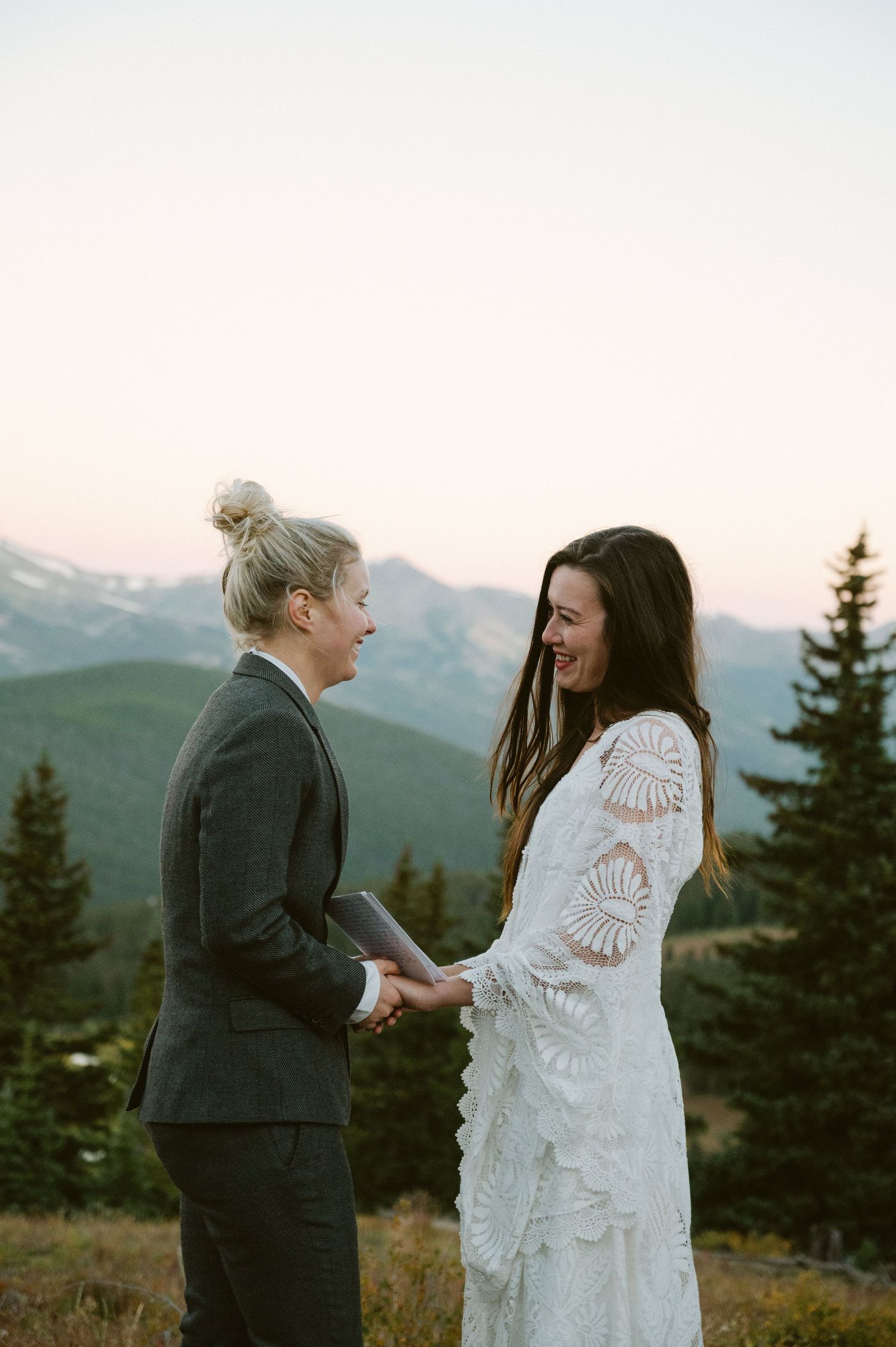 Two people laughing together on a mountaintop during sunset with evergreen trees in background.