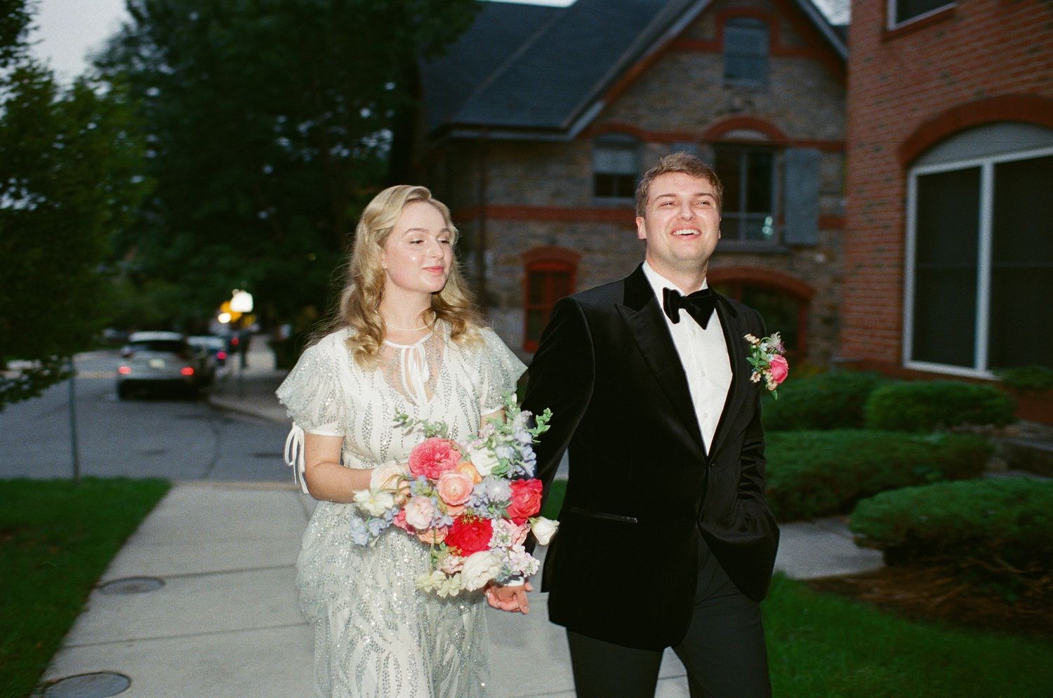 A couple in formal attire poses outside a brick building with bouquet.