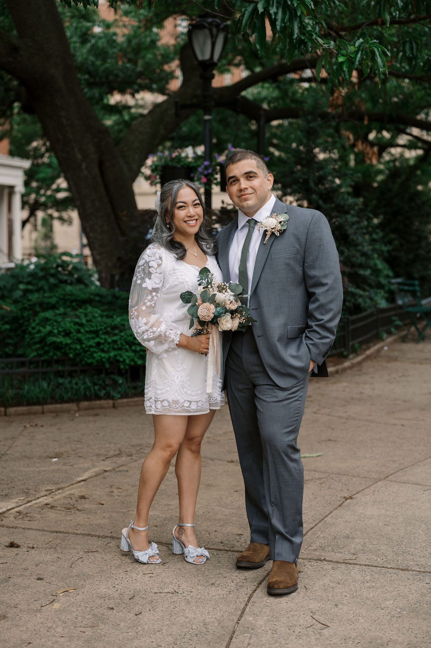 A couple stands together on a tree-lined path, the bride wearing a short lace dress and the groom in a grey suit.