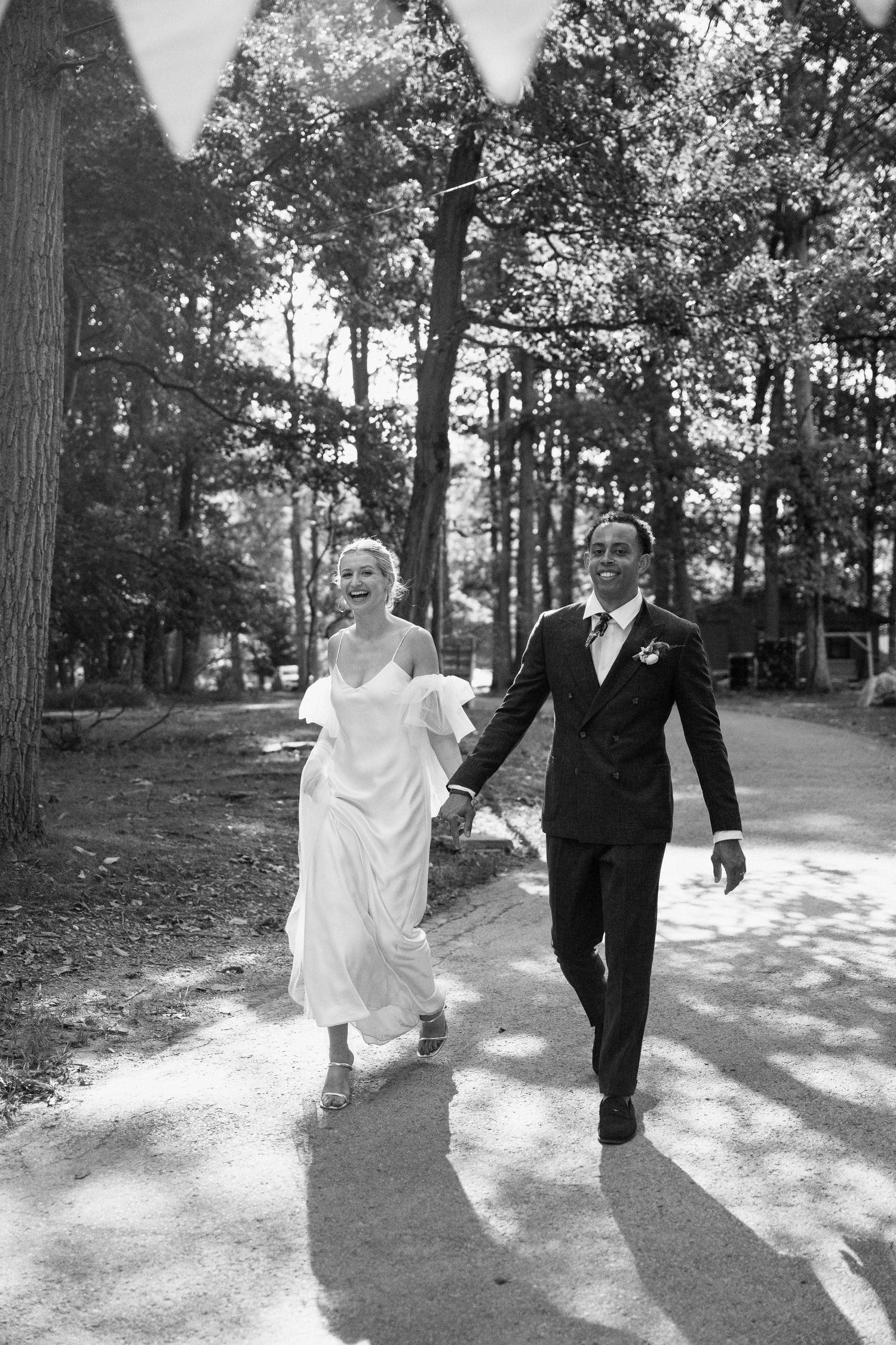 A couple walks joyfully down a tree-lined path after their wedding ceremony.