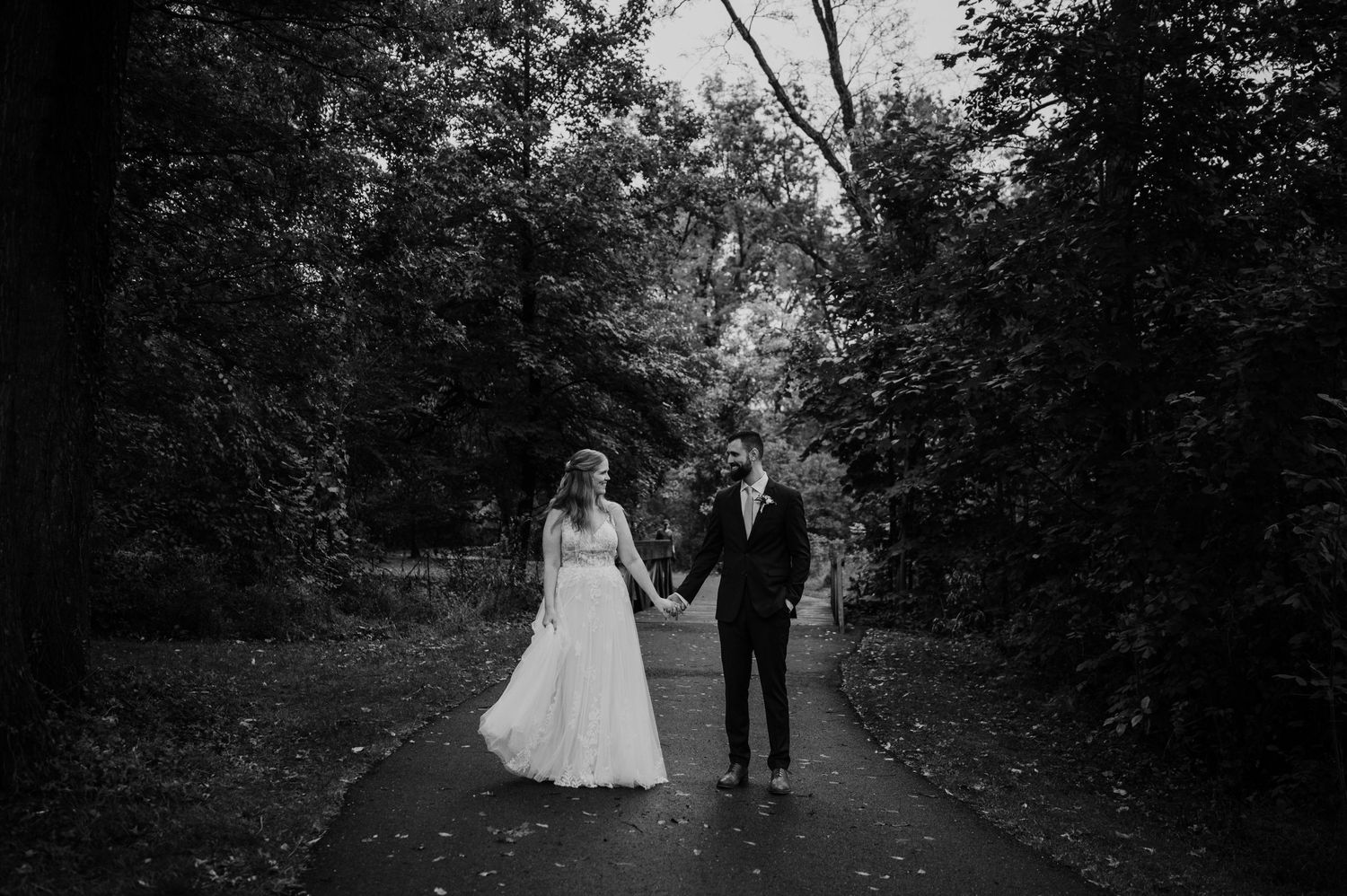 A black and white photo of newlyweds walking hand in hand down a wooded path.