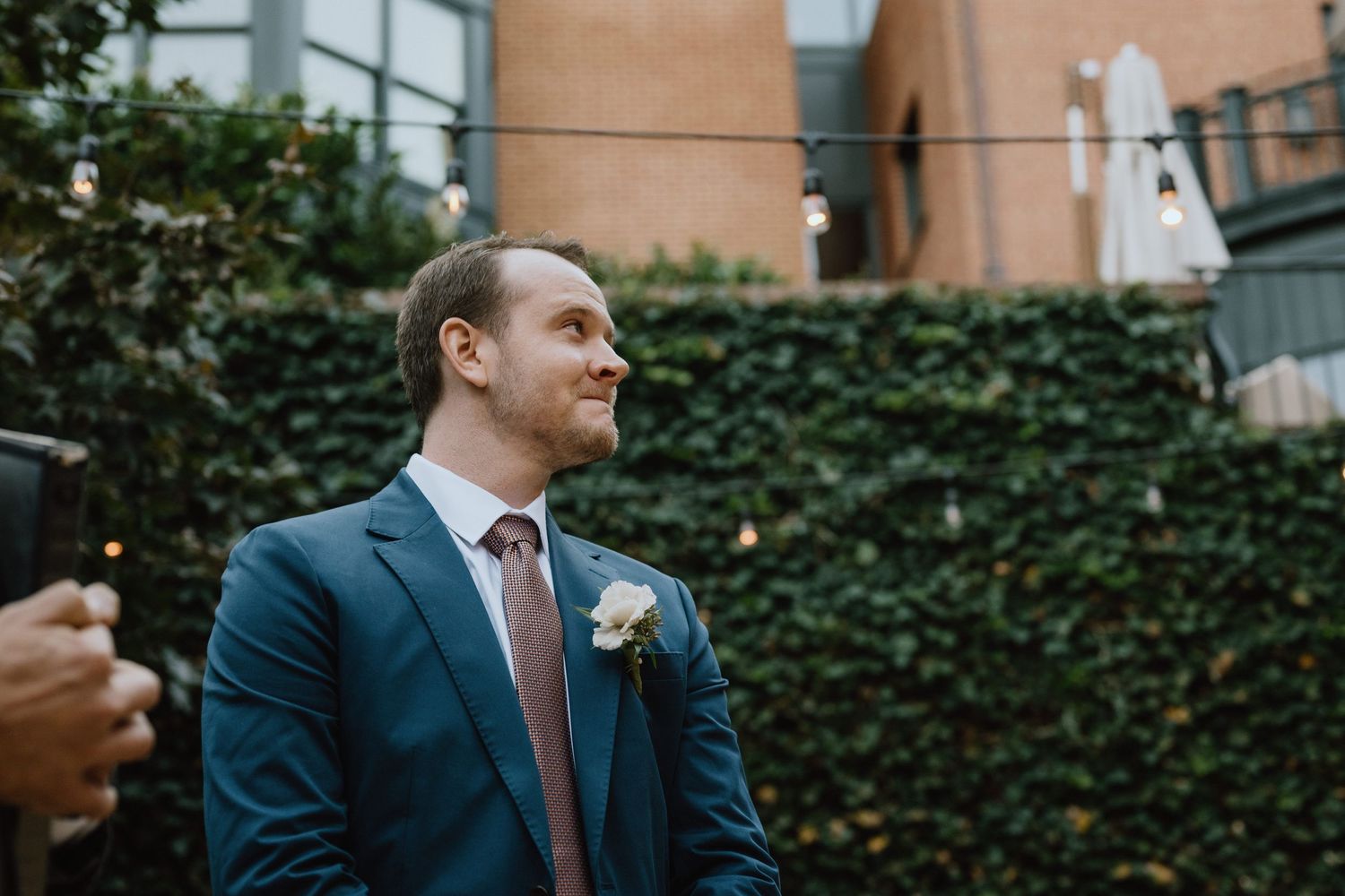 A suited person with boutonniere smiling at an outdoor event with ivy wall backdrop.
