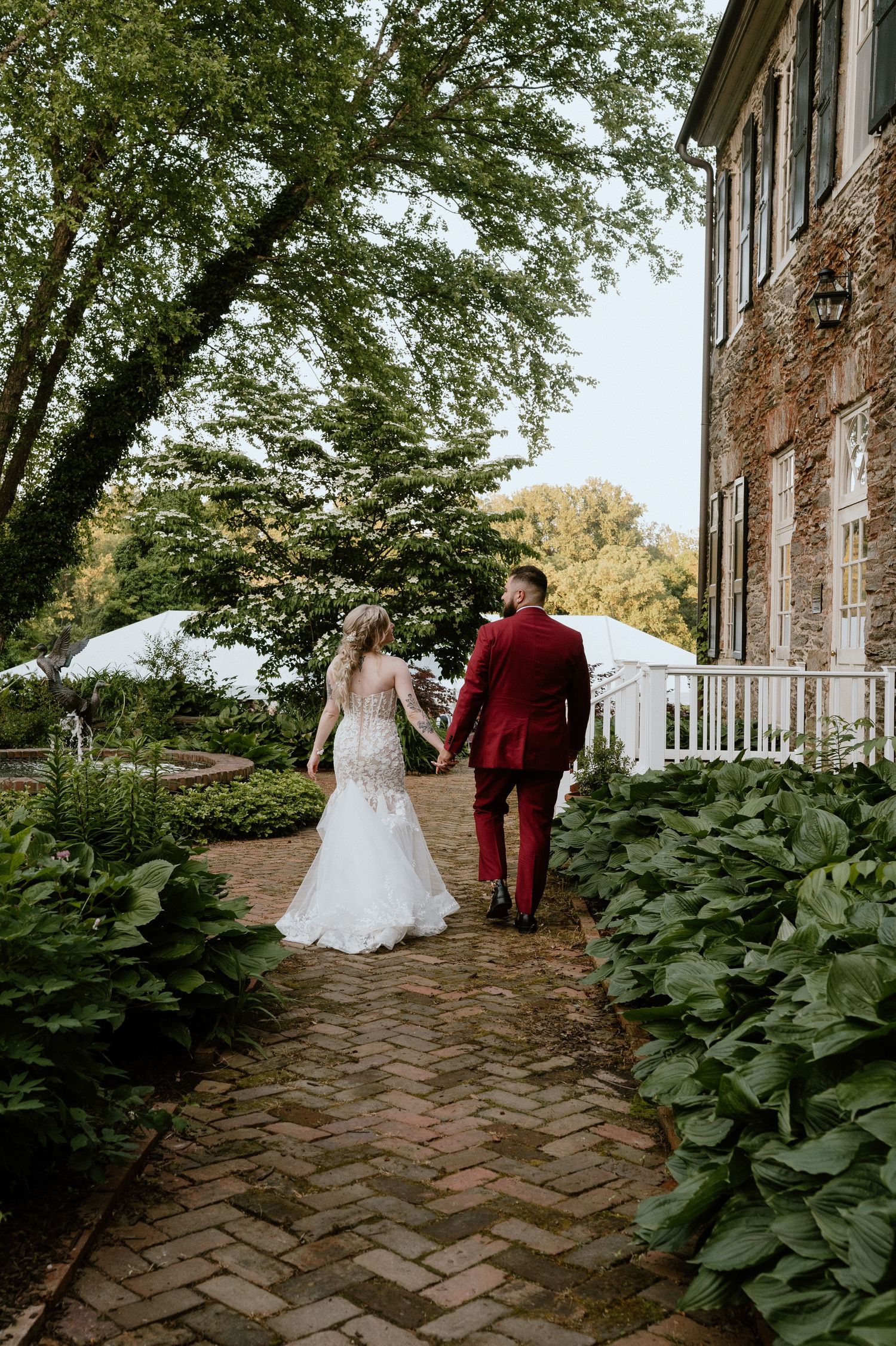 A couple walks along a brick path beside a historic stone building with lush greenery and white tents in the background.