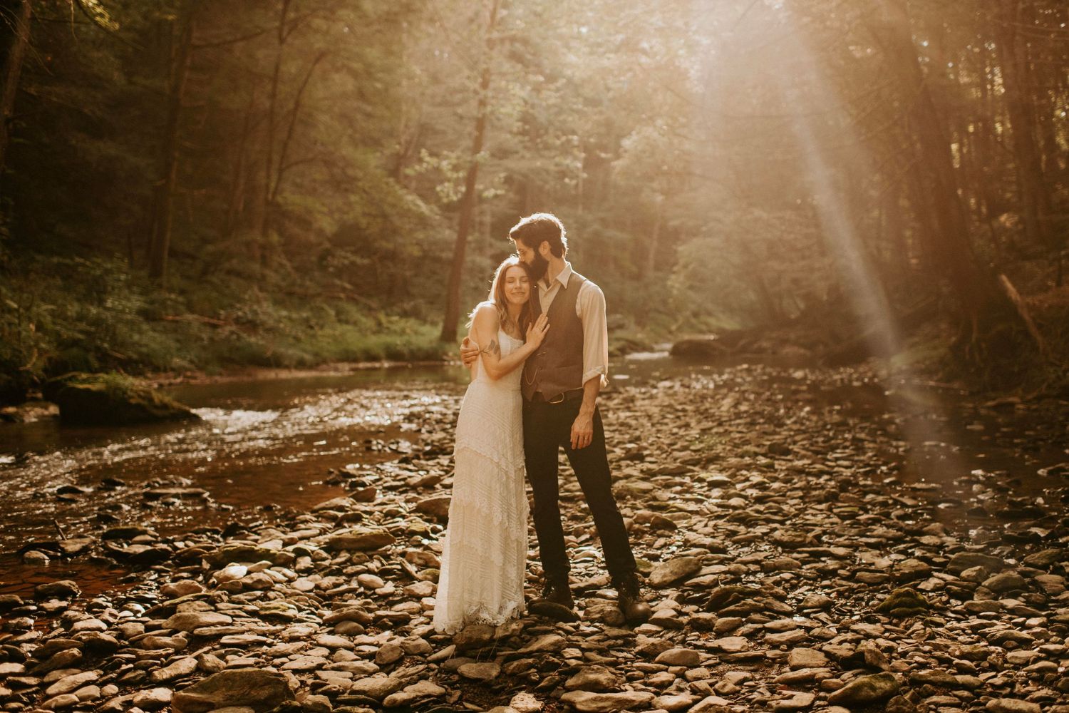 A couple embraces in sunlit forest with golden light streaming through trees onto fallen leaves.