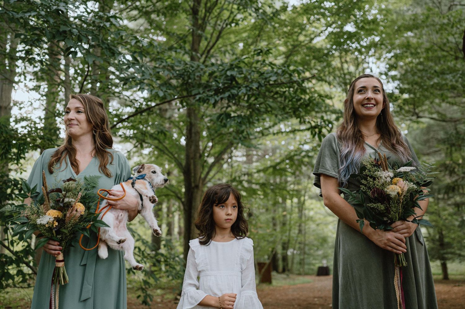 Three people holding bouquets of flowers stand together in a lush forest setting during a wedding ceremony.