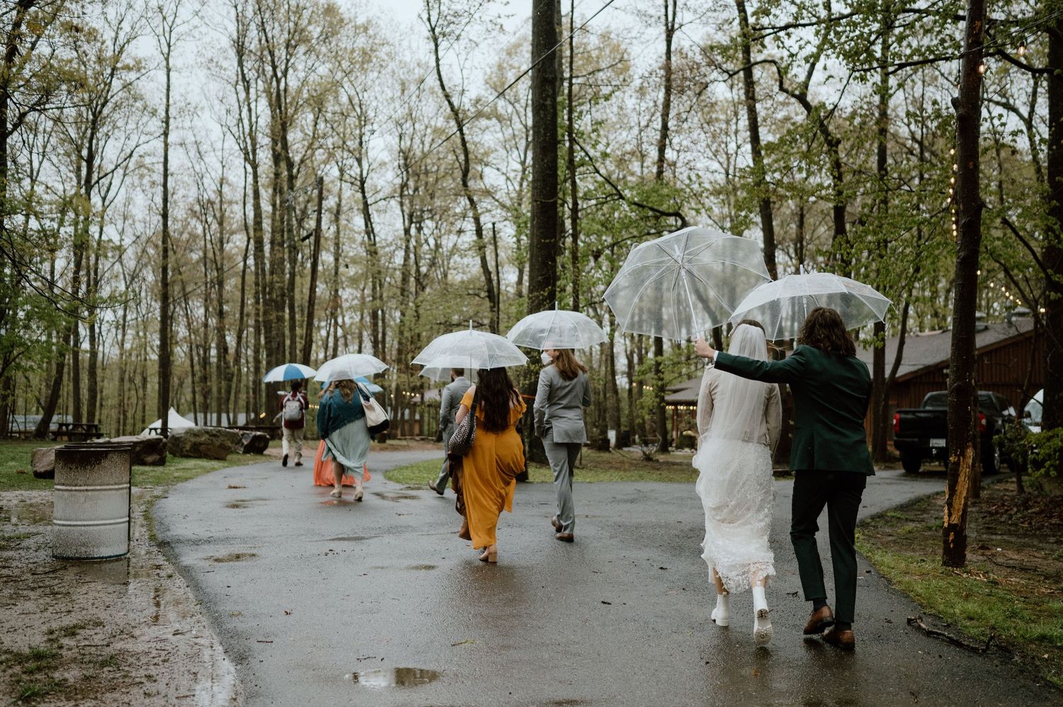 Wedding party walking with umbrellas on wet pathway through wooded area.