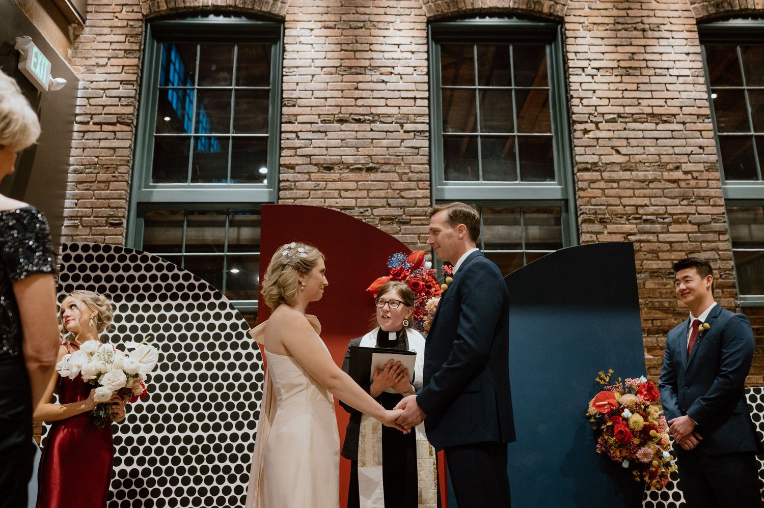 A wedding ceremony takes place in front of a red and navy geometric backdrop against exposed brick walls.
