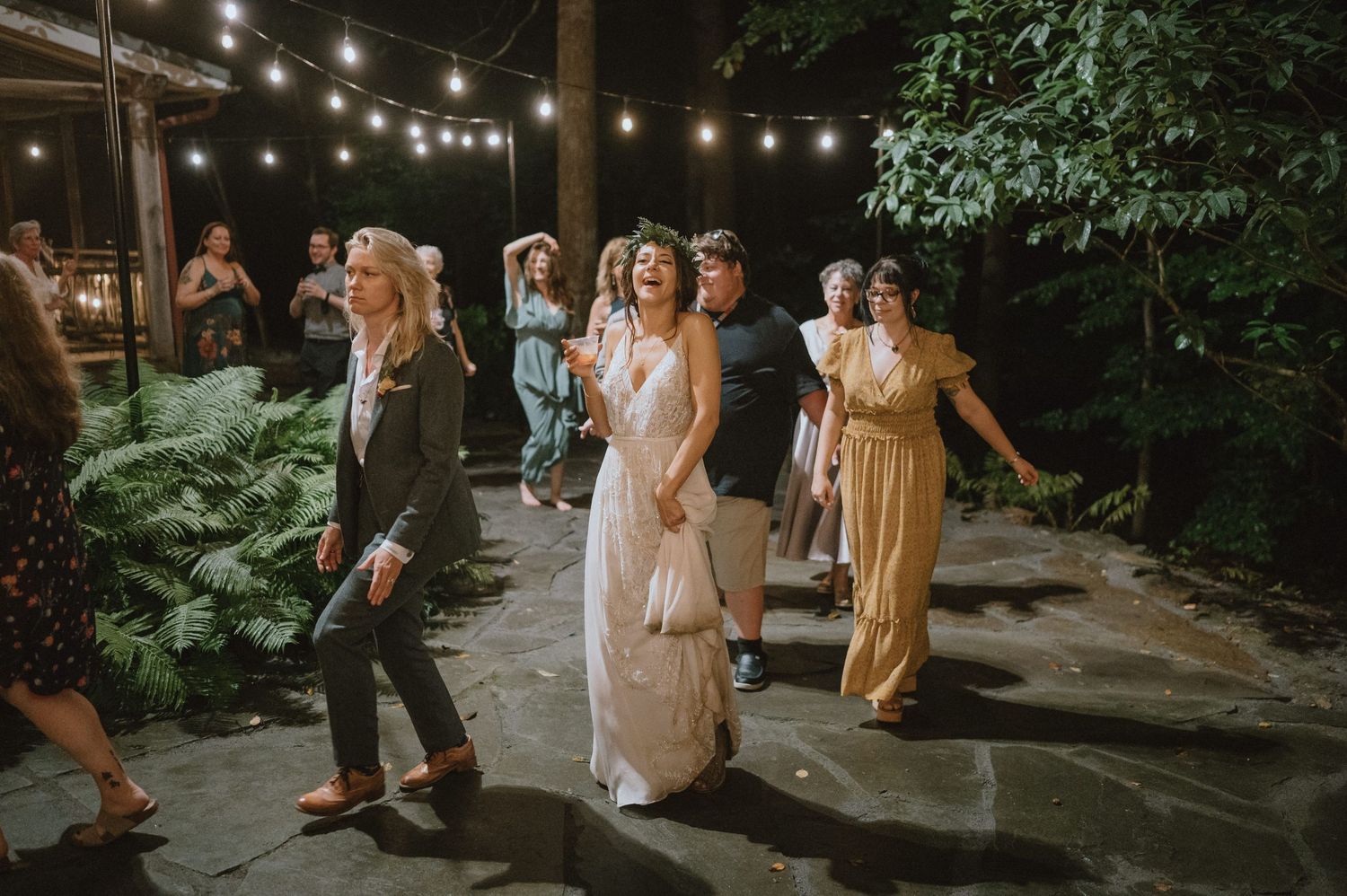 Wedding guests dance under outdoor string lights on a stone patio surrounded by lush greenery.