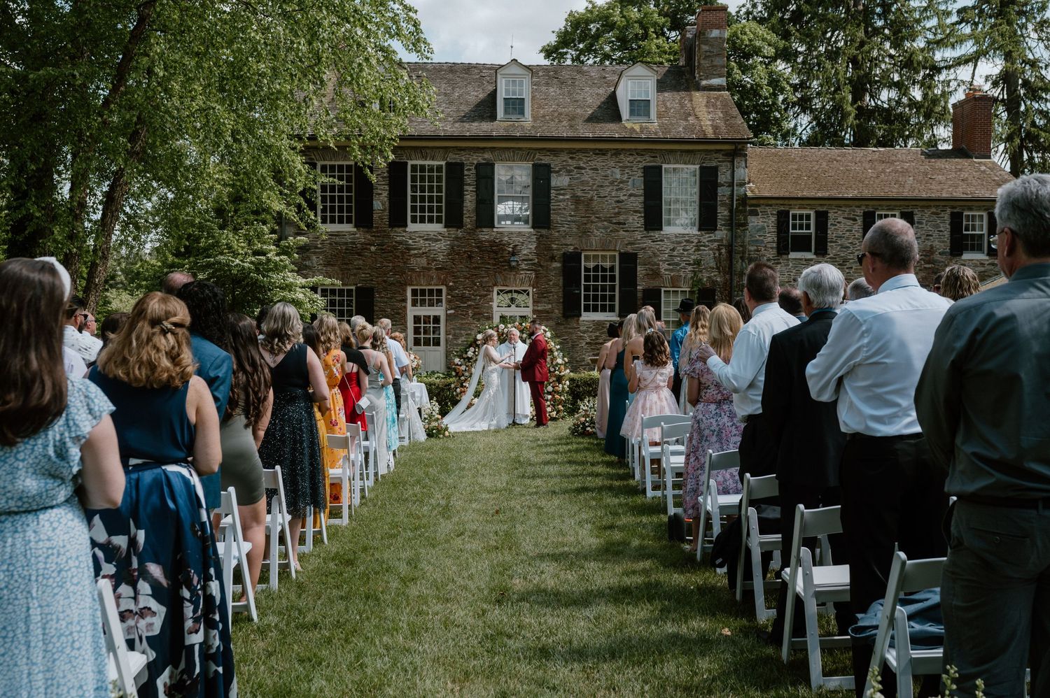 Outdoor wedding ceremony at historic stone mansion with guests seated on white chairs.