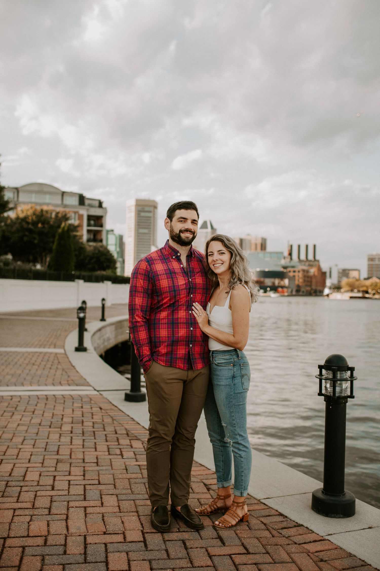 Couple standing on brick waterfront with city skyline in background.