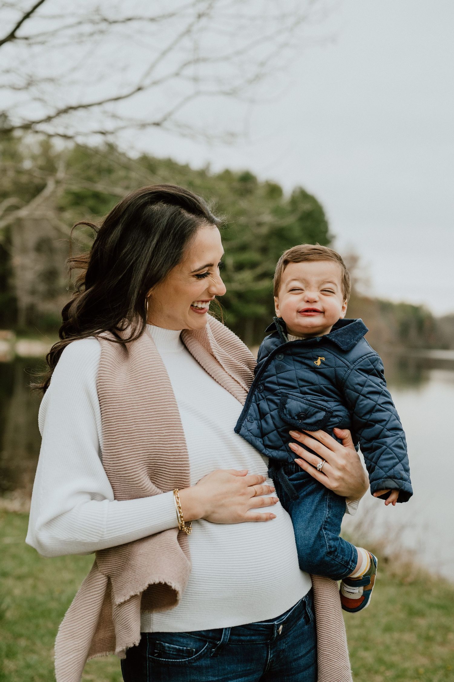 Expecting parent in white sweater holds toddler by lakeside on autumn day.