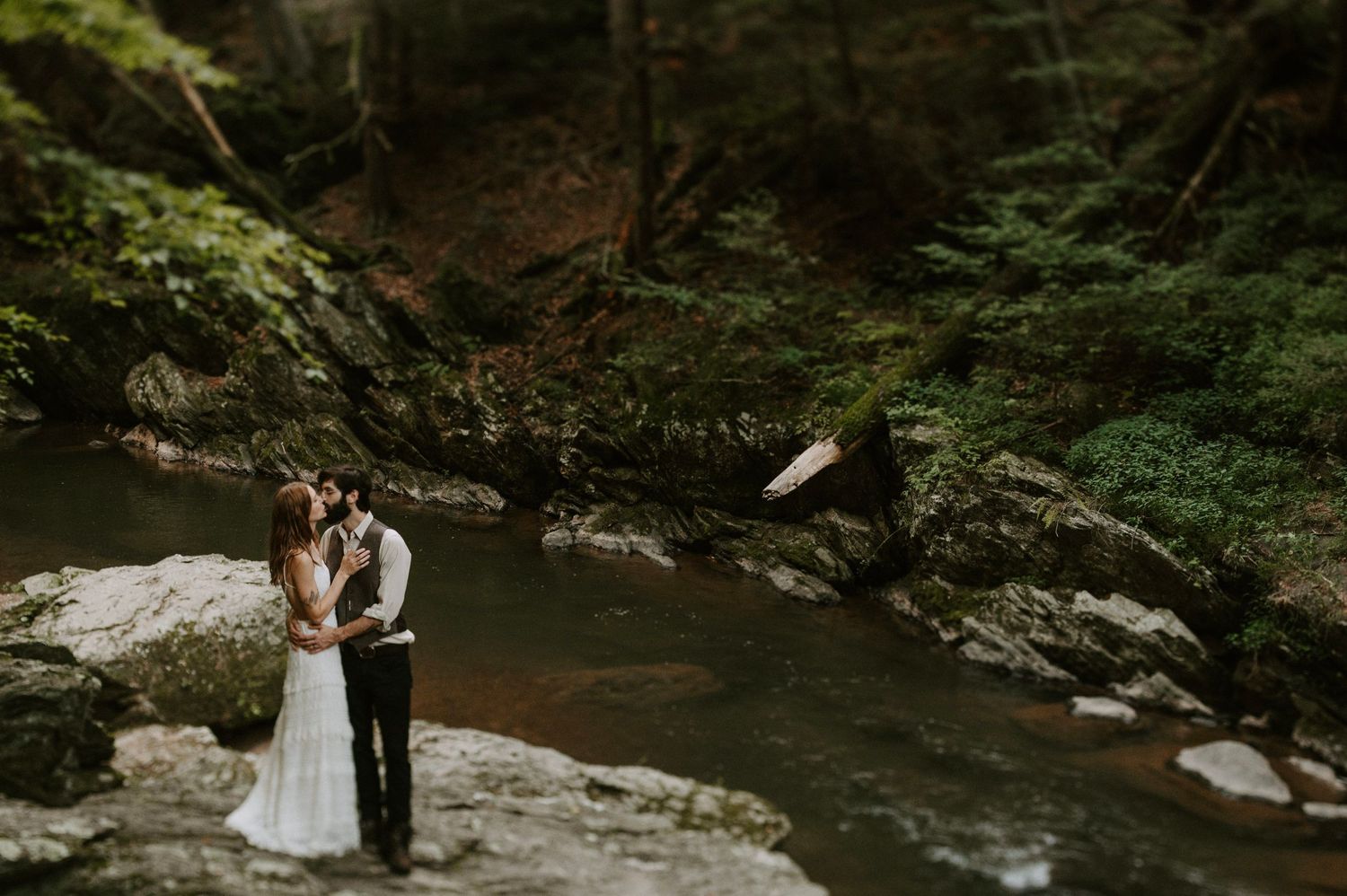 A couple stands intimately on rocky outcrop overlooking scenic forest stream in moody natural setting.