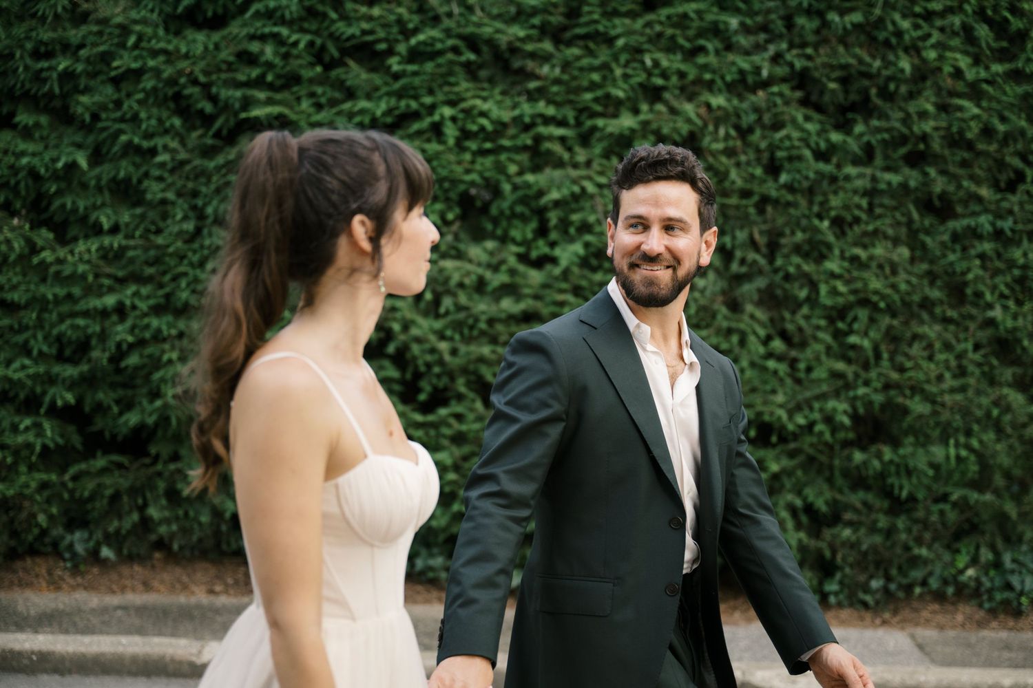 A couple in formal wedding attire laughs together while walking along a lush garden path on their wedding day.