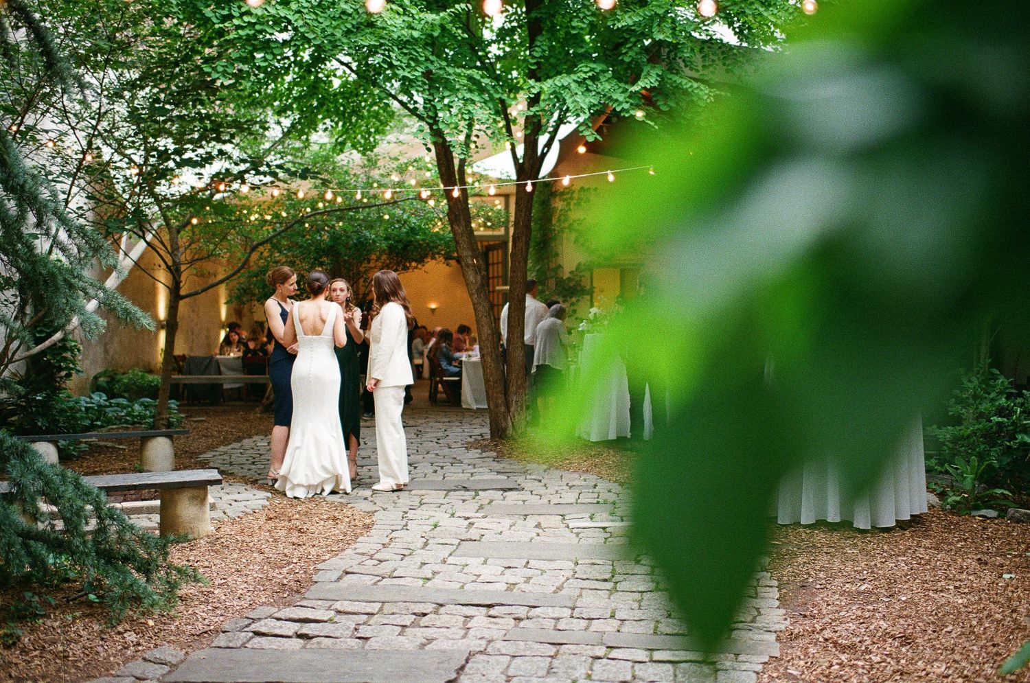 A couple in white dresses walks along a brick path surrounded by lush garden foliage and warm ambient lighting.