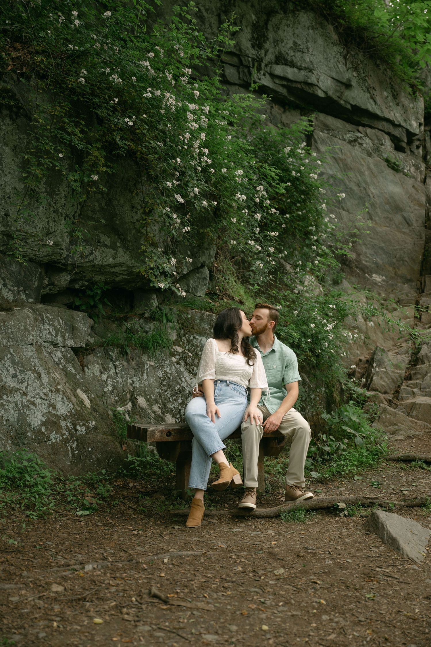 Couple sharing a kiss on a rustic wooden bench surrounded by lush greenery and rocky cliffs.