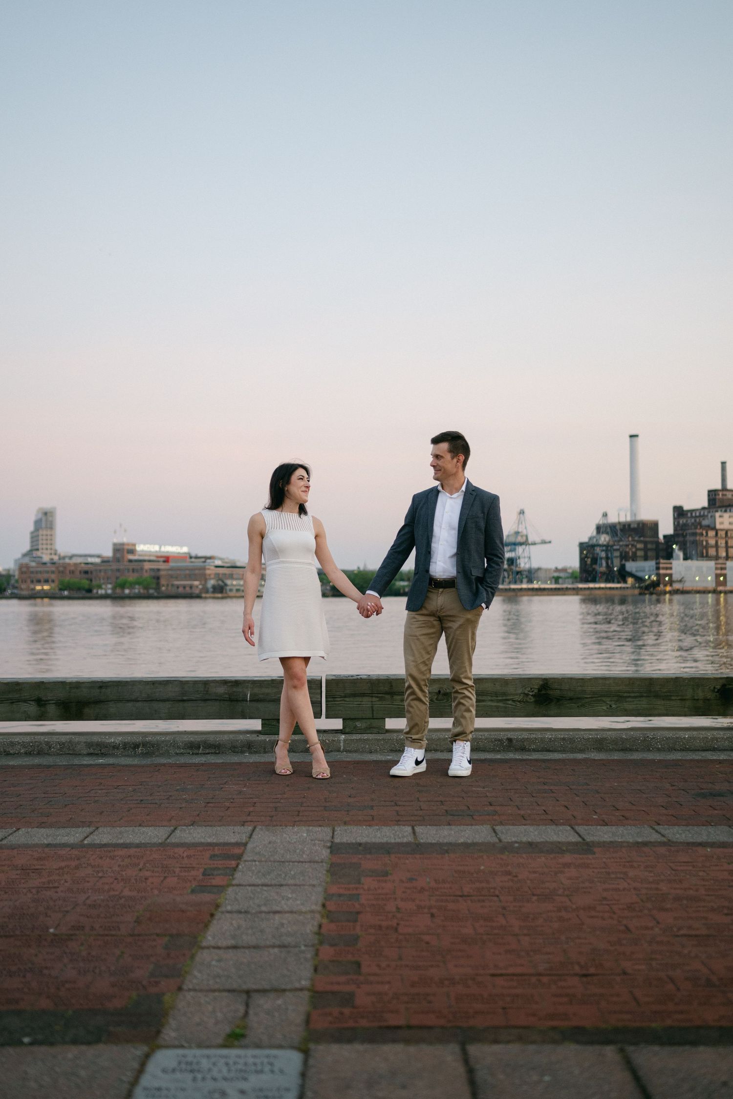 Couple holding hands on a waterfront promenade at dusk, woman in white dress, man in blazer and khakis.