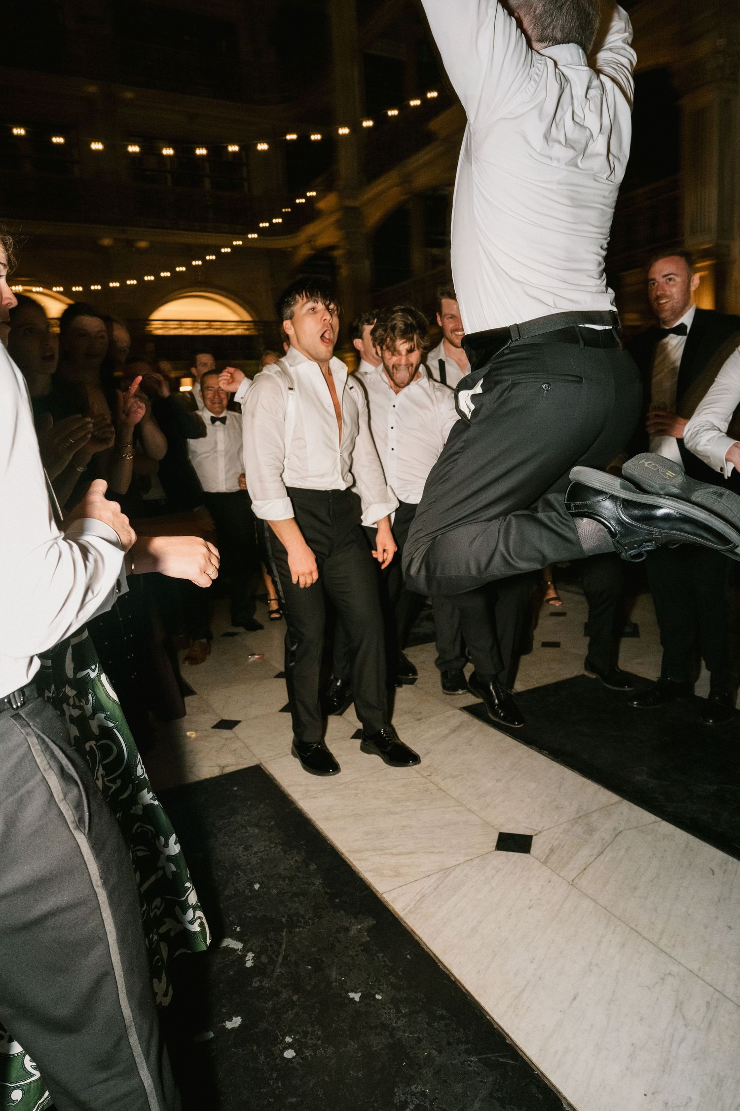 Wedding reception guests dancing and jumping with string lights visible in the background.