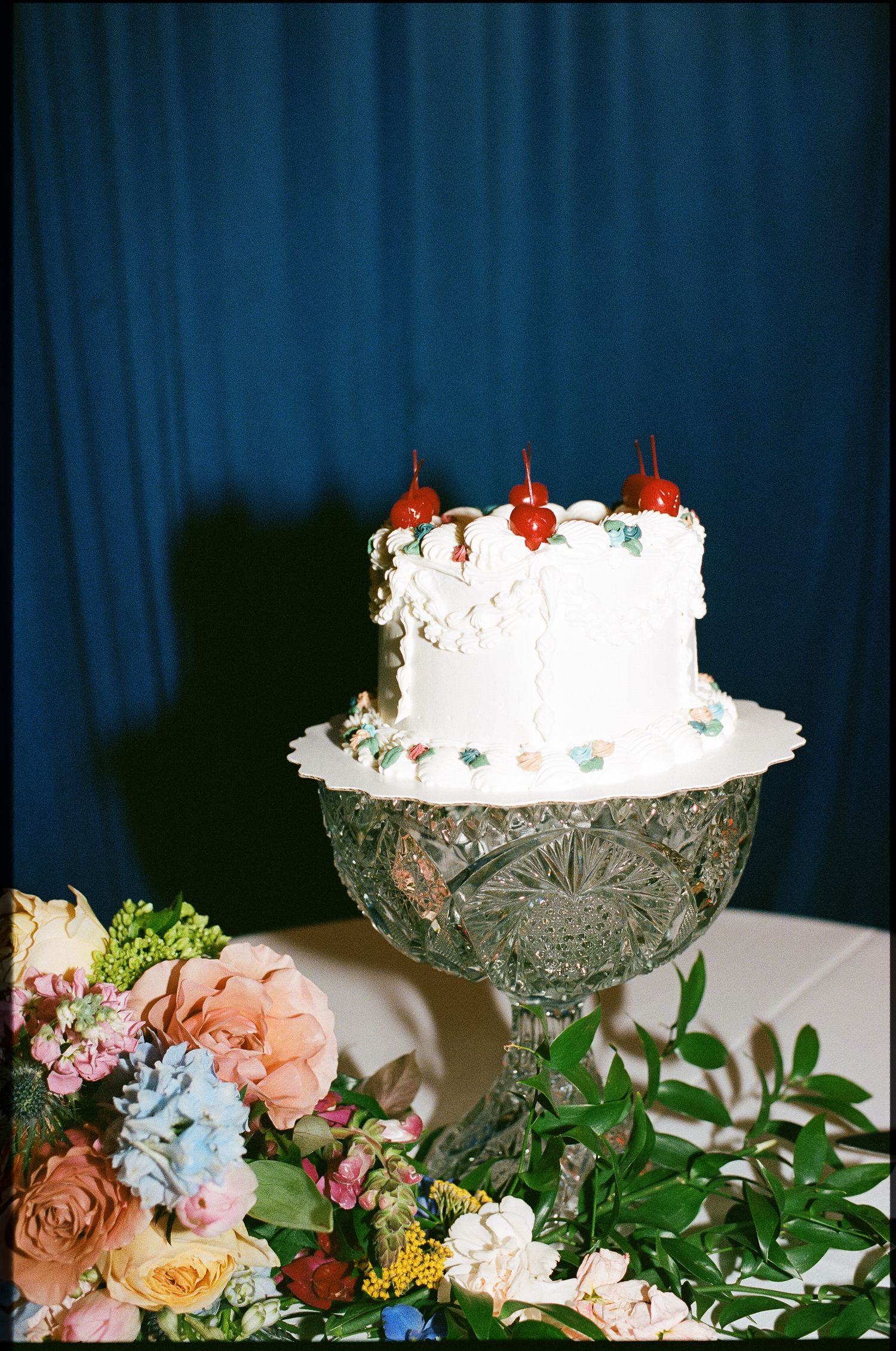 White frosted cake with cherry toppers on crystal stand surrounded by colorful floral arrangement against blue backdrop.