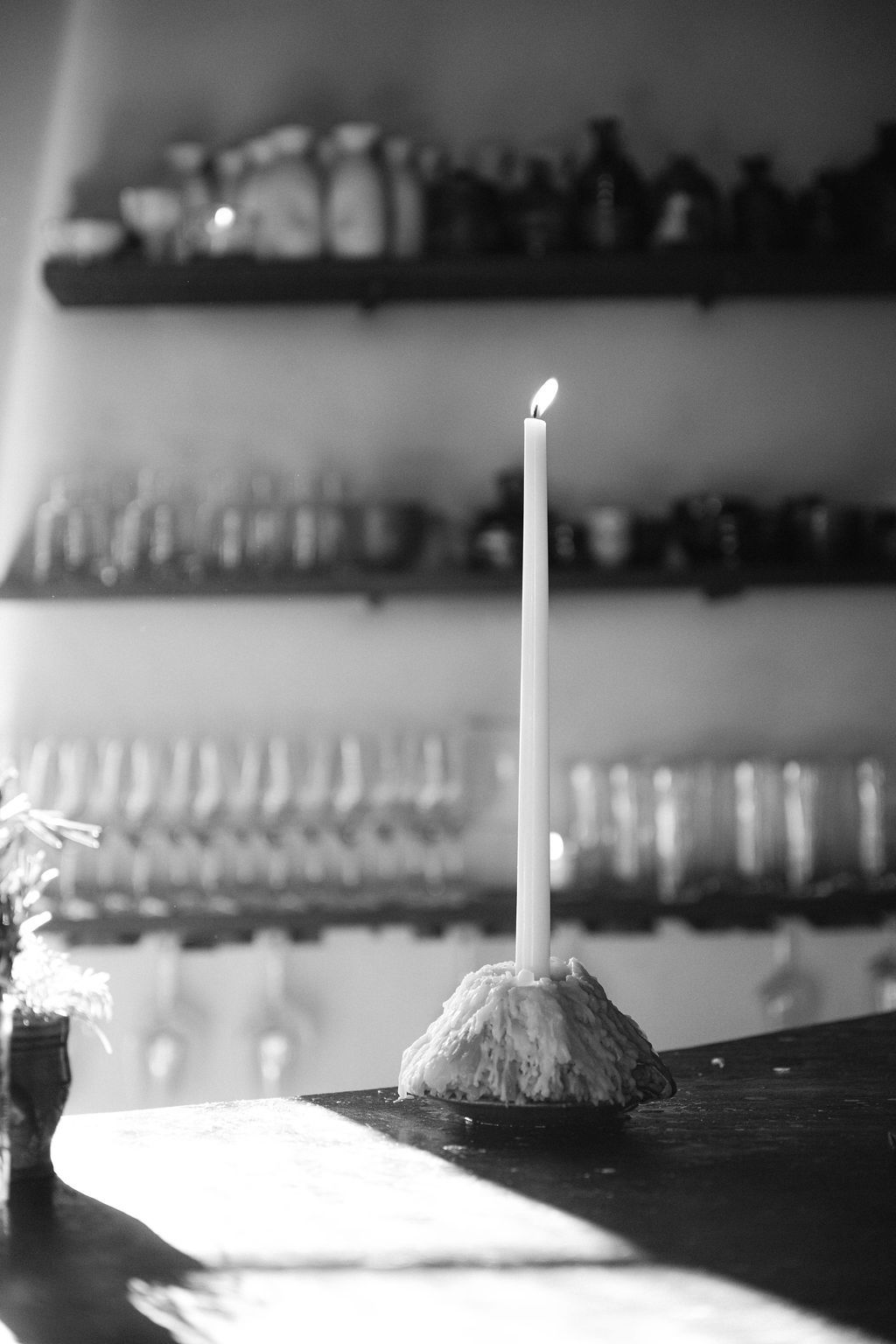 A single lit white candle on a rustic surface with blurred glassware in the background.