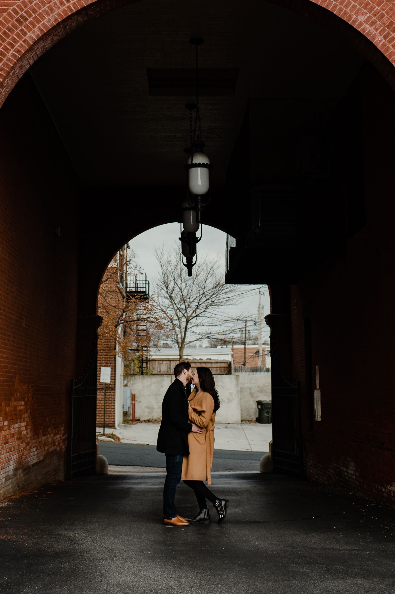 Couple sharing a romantic moment under a brick archway with hanging lanterns in an urban setting.