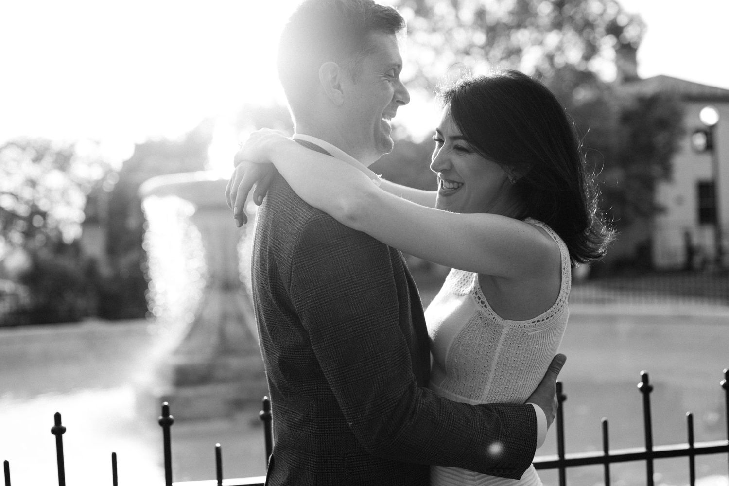 Black and white photo of a couple embracing and smiling outdoors, with sunlight glowing behind them.