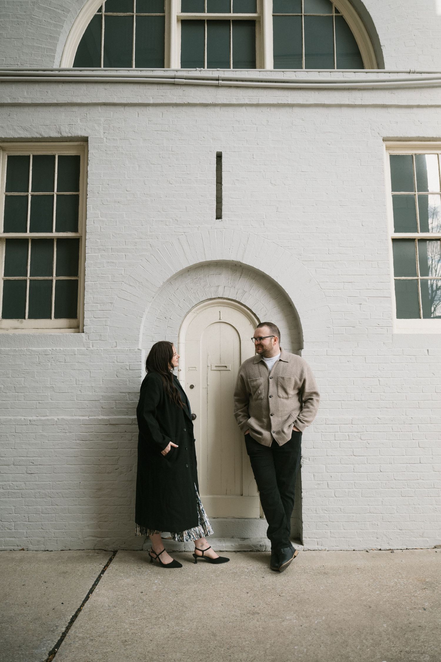 Couple standing and talking in front of a white brick building with an arched door.