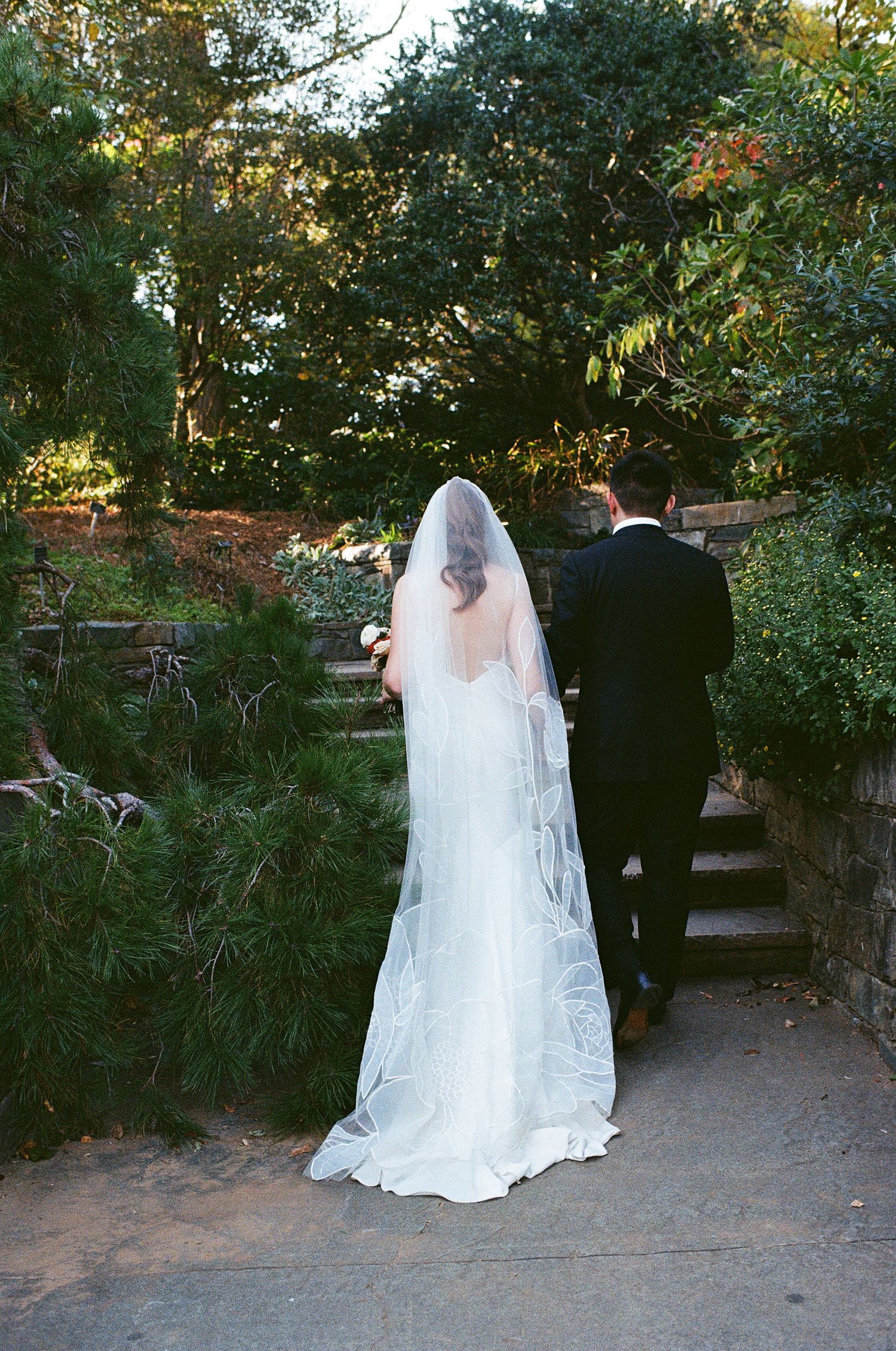 Wedding couple walking through a garden path surrounded by lush greenery at sunset.