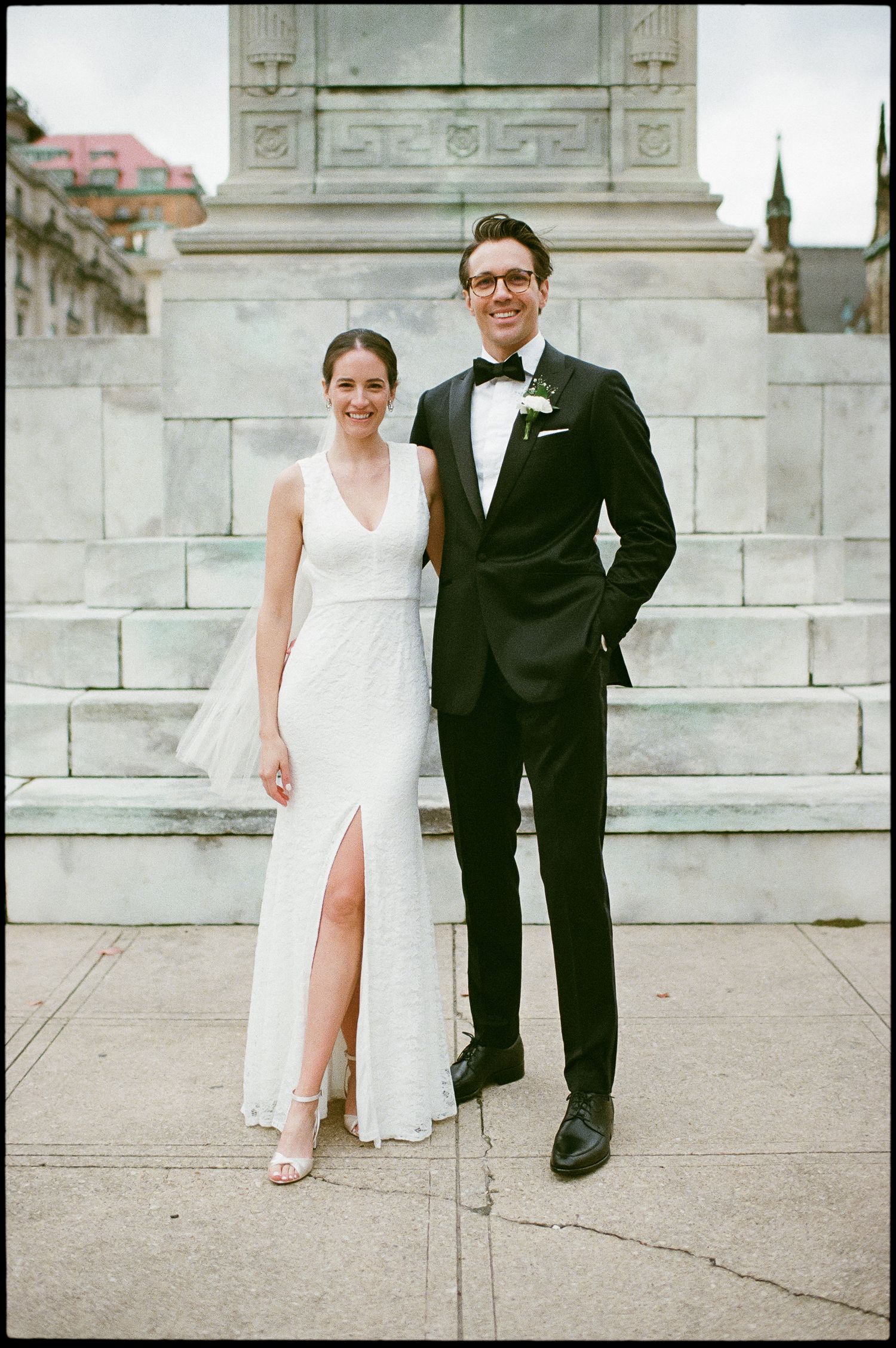 A bride in a white gown with a slit and groom in black tuxedo pose together on stone steps in an urban setting.