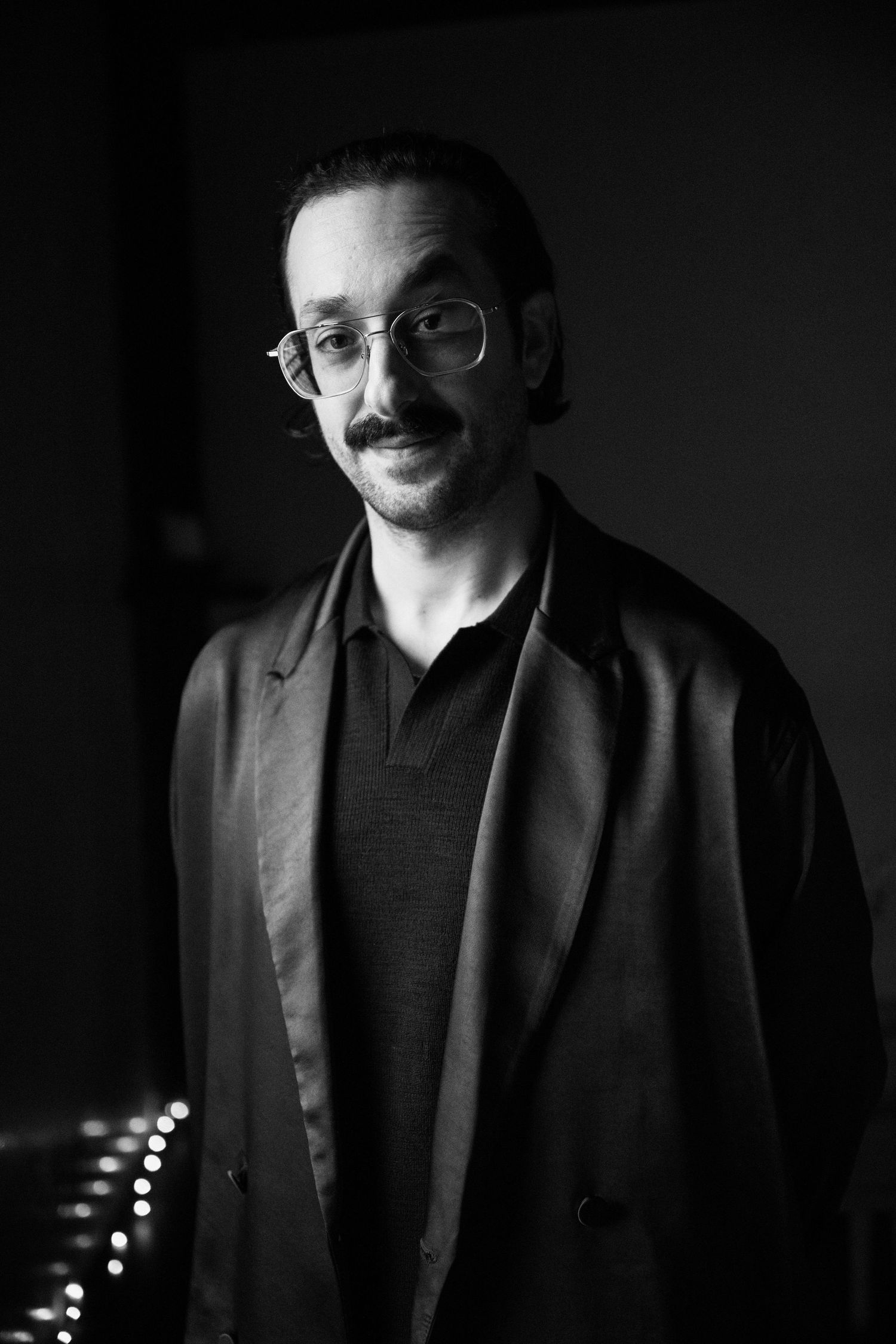 Portrait of a man with glasses and mustache in black clothing against dark background at Fadensonnen in Baltimore