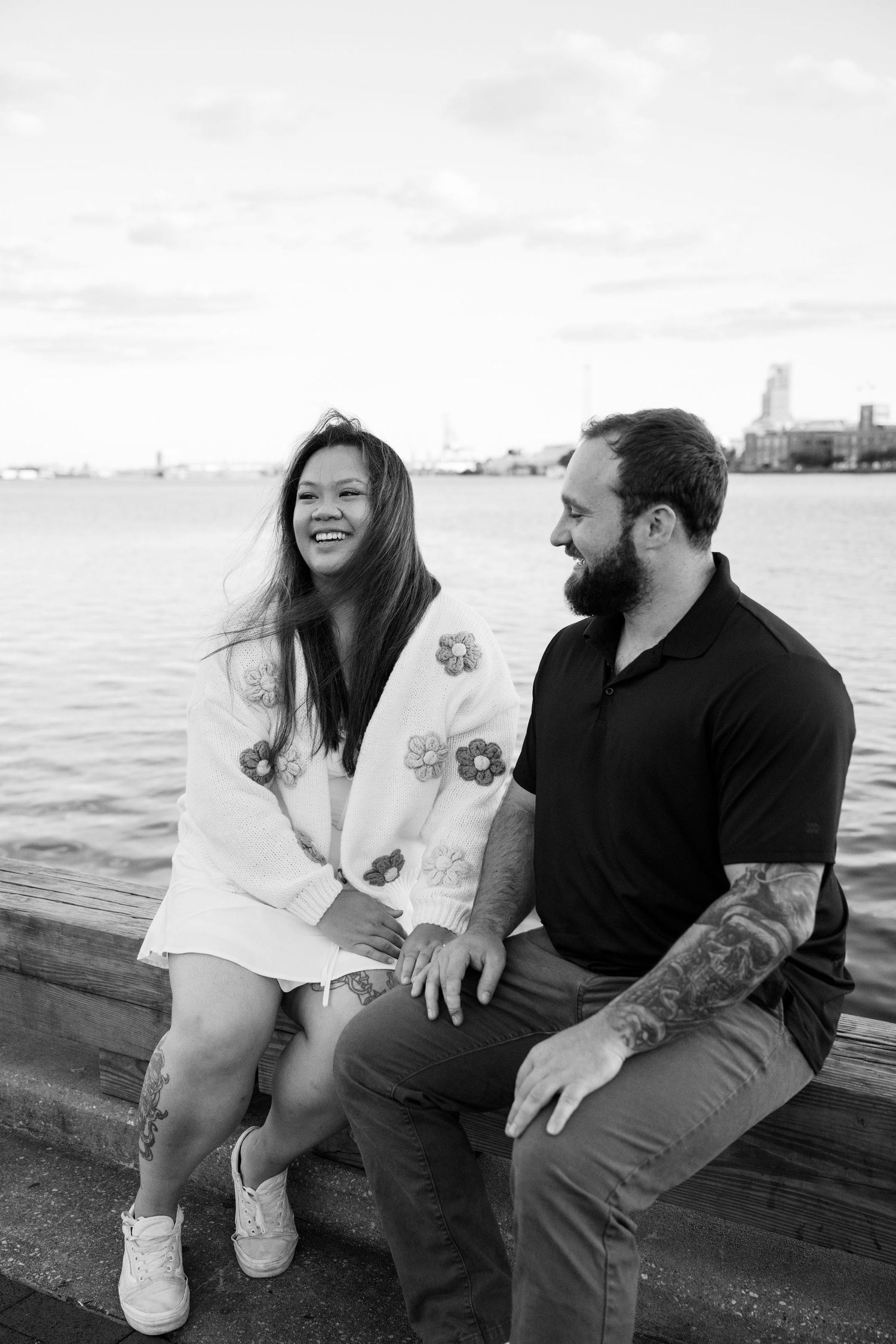 A couple sits on a waterfront pier sharing a candid laugh together in black and white.