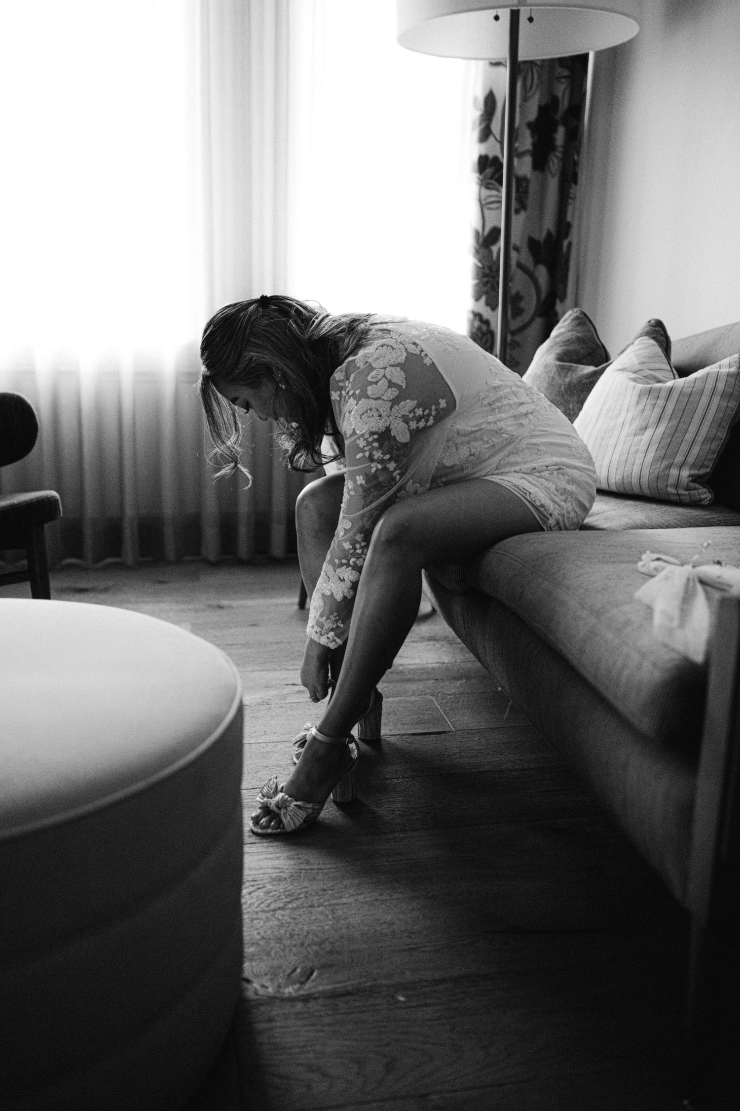 A person in a white dress sits on a couch while putting on shoes in a modern room.