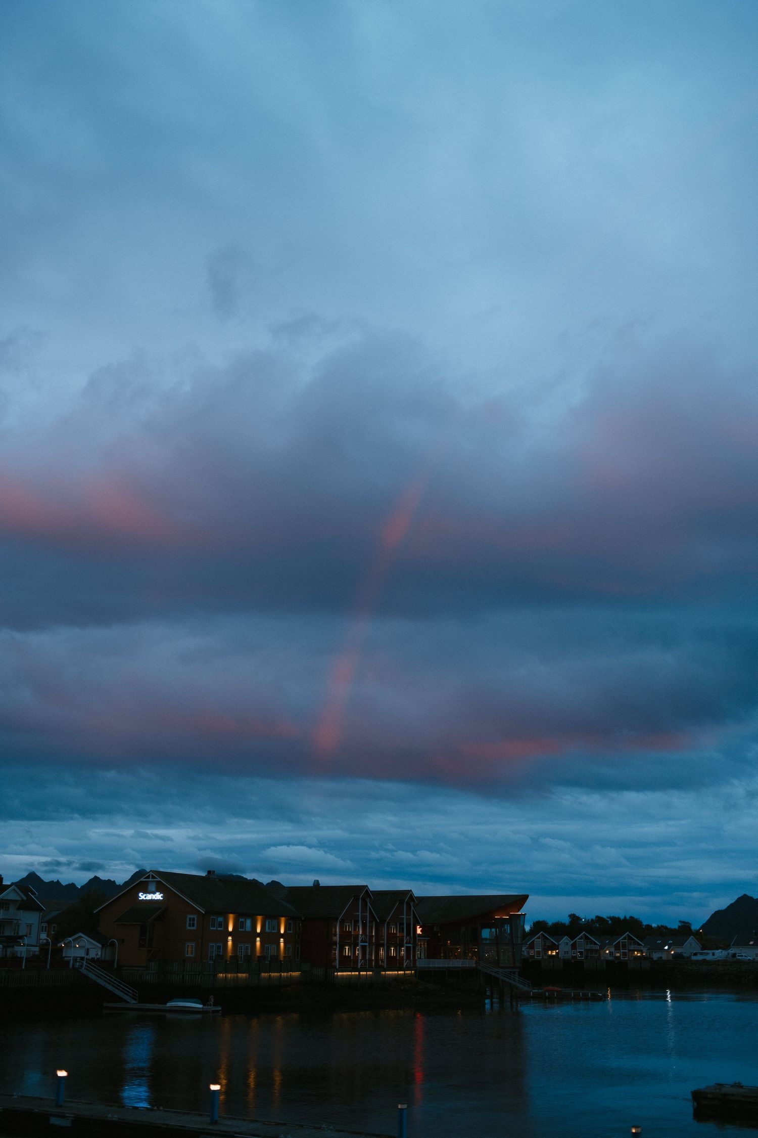 Pink sunset clouds and rainbow over waterfront buildings reflected in calm harbor waters.