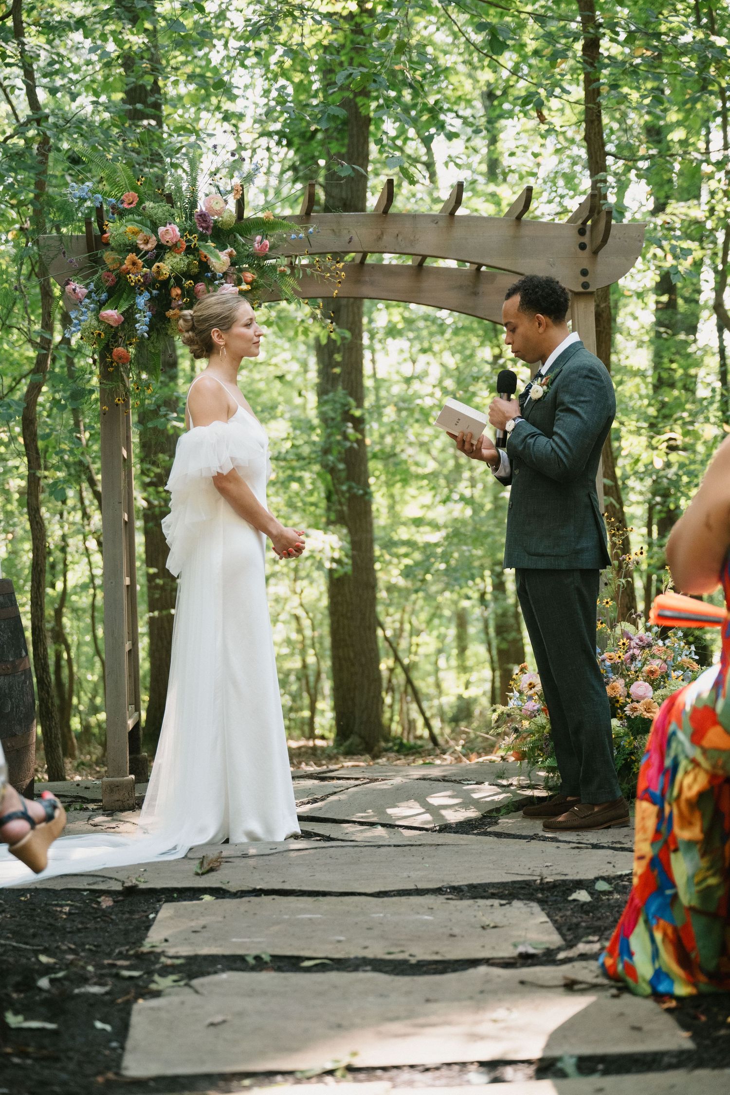A couple exchanges vows under a flower-adorned wooden arch in a forest setting.