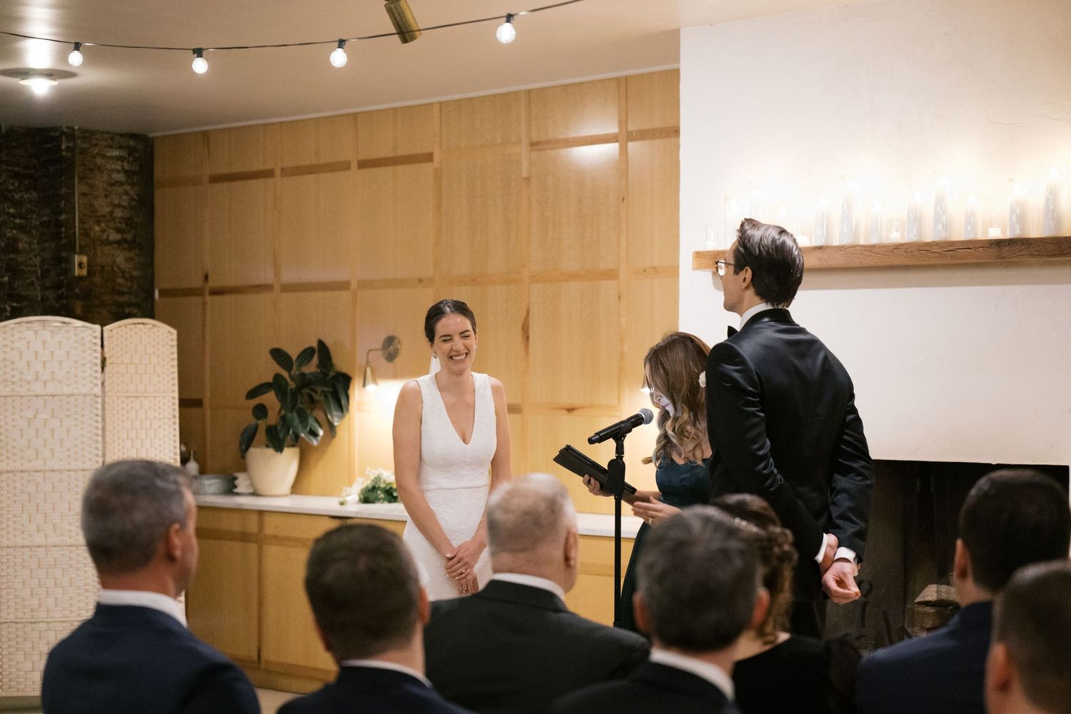 Wedding ceremony with a minister speaking to a couple at the front of a modern venue with string lights and guests looking on.