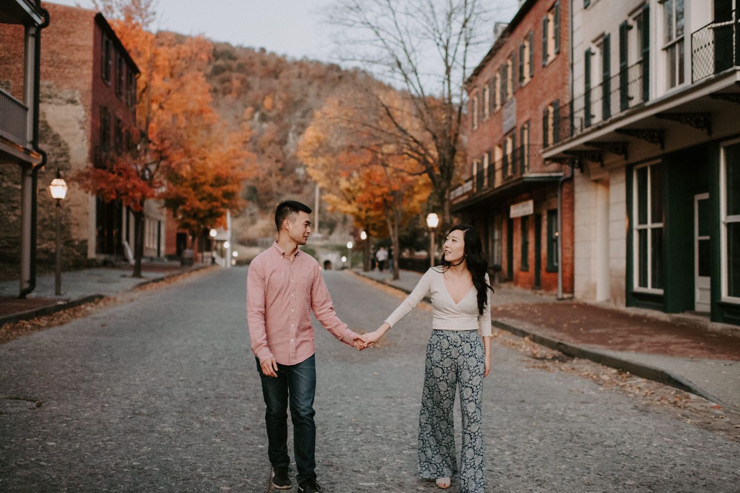 A couple holds hands while walking down a historic brick street with autumn foliage.